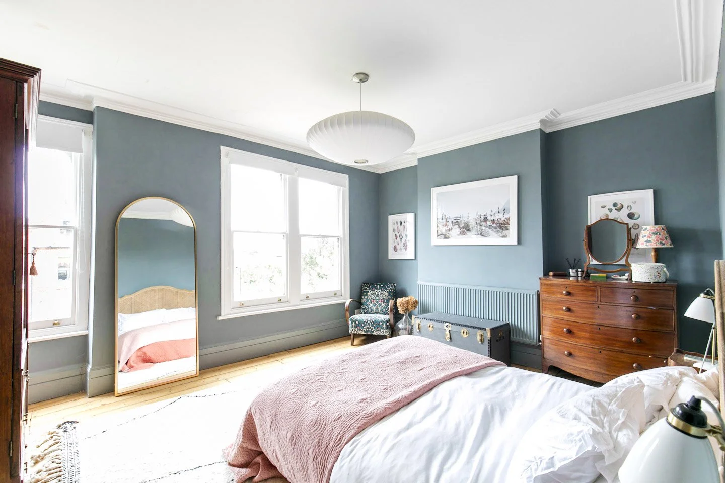 A bedroom with teal walls, large windows, a bed with white and pink bedding, a wooden dresser with a mirror, a patterned armchair, a mirrored full-length wardrobe, framed wall art, and a hanging ceiling light.