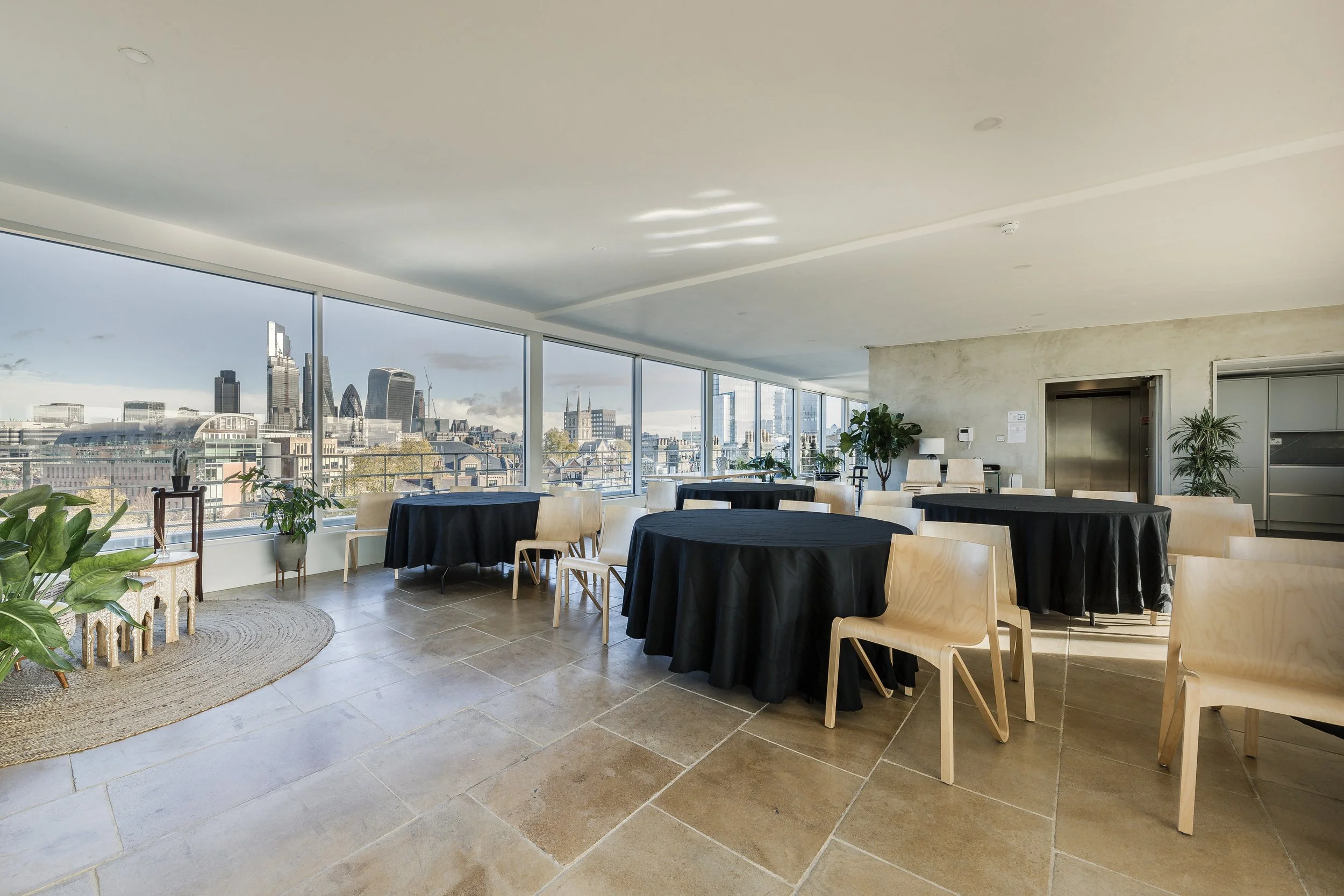 An interior space with large windows showing a cityscape view, round tables with black tablecloths, wooden chairs, and potted plants, inside a modern building.