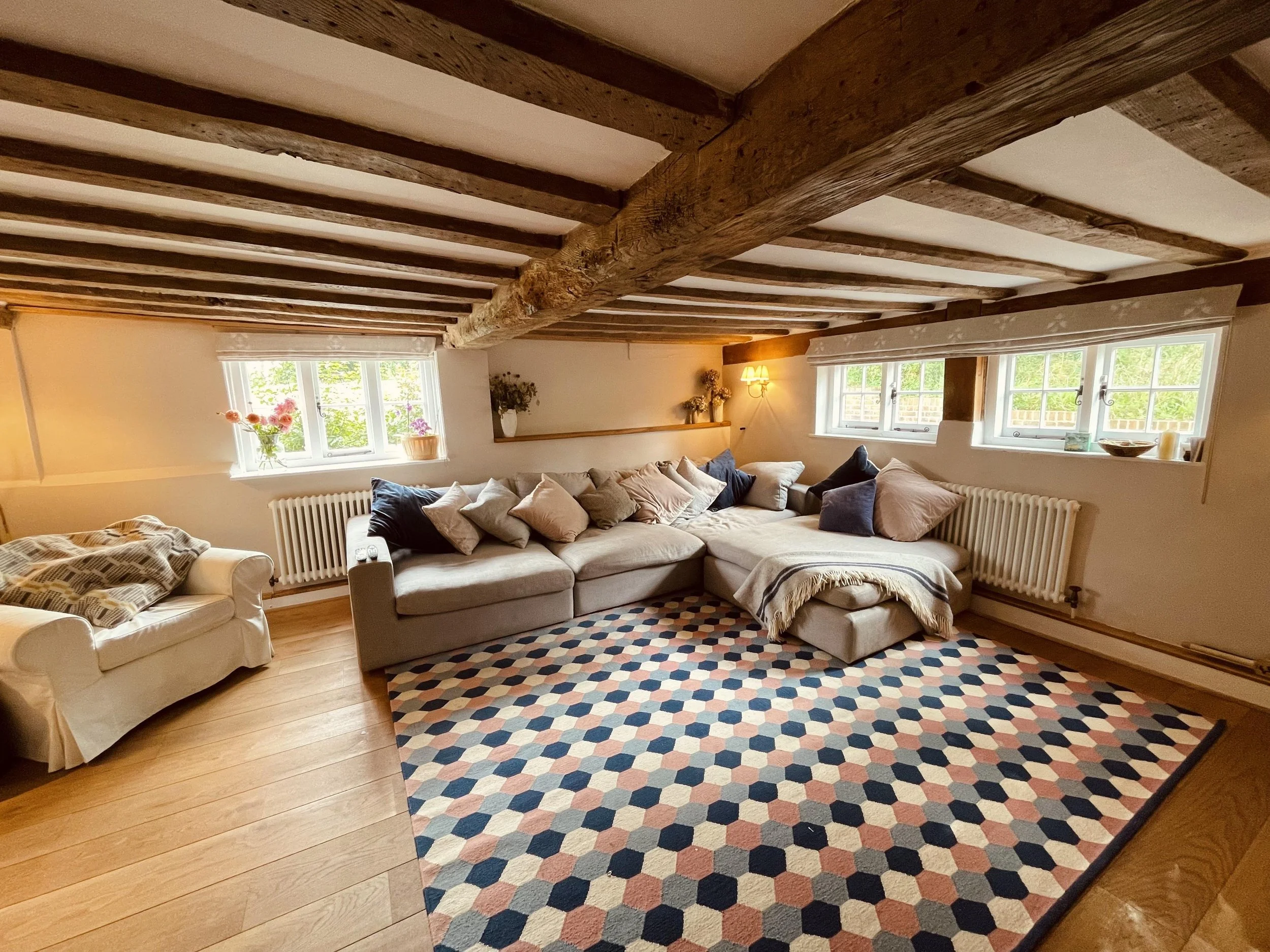 Living room with a sofa, armchair, colorful geometric patterned rug, and wooden ceiling beams. There are windows with white curtains and a table with flowers.