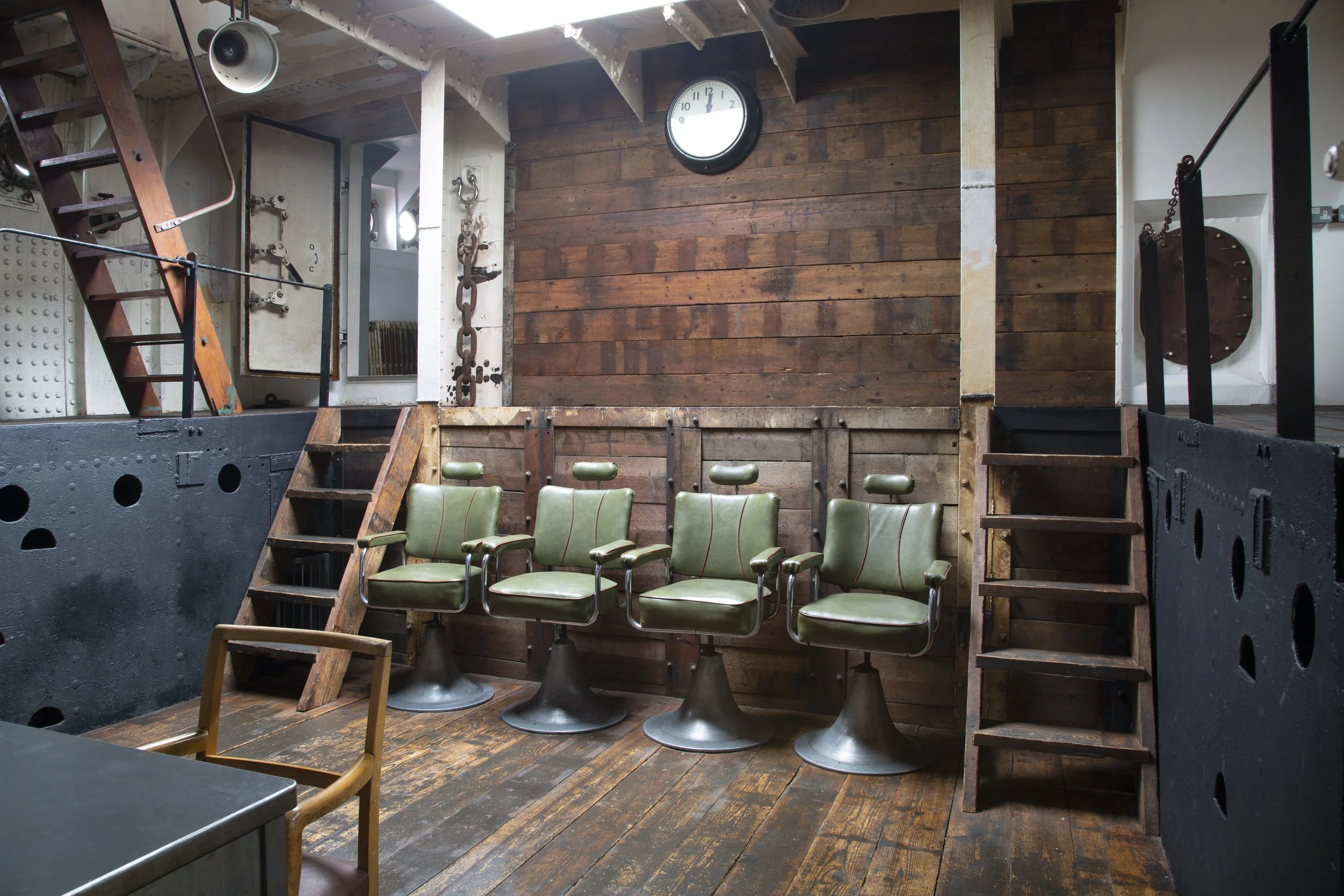 Interior of a rustic room with four green swivel chairs, wooden floors, and wooden walls, featuring a clock and industrial décor.