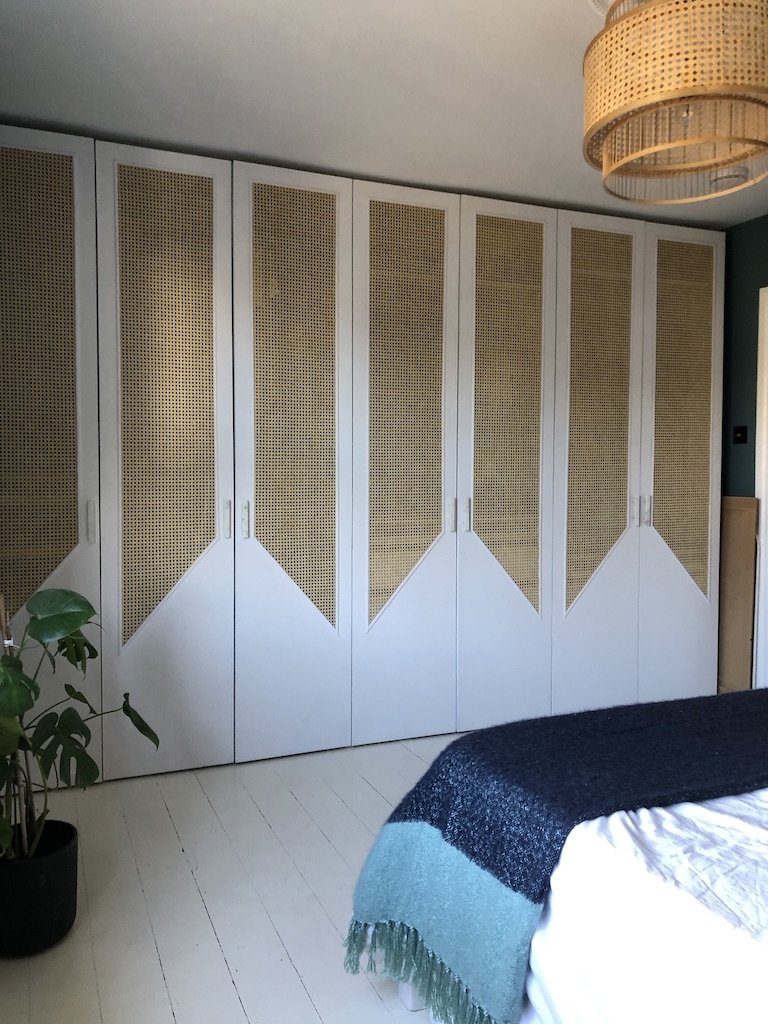 Bedroom with built-in closet doors featuring a combination of white and woven rattan panels, a potted plant in the corner, part of a bed with a dark blanket and light bedding, and a woven ceiling light fixture.
