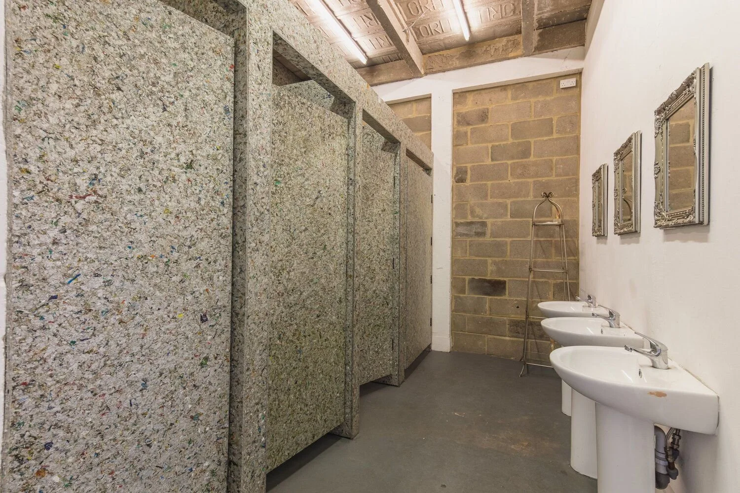Public restroom with three sinks, a wall-mounted mirror, and colorful bathroom stalls in an unfinished space with exposed brick and plaster walls.