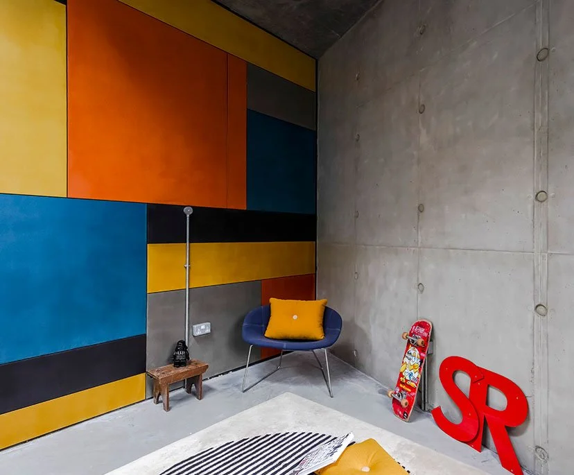 A corner of a room with a colorful, geometric wall design, concrete wall, a small wooden side table with a black lantern, a black armchair with a yellow pillow, a skateboard propped against the wall, and large red letters 'SR' on the floor.