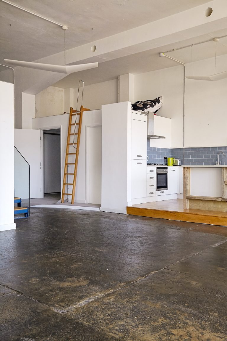 Empty room with concrete floor, white walls, and an open kitchen with white cabinets, a gray tile backsplash, and a wooden step. A yellow kettle is on the counter and a wooden ladder leans against a wall.