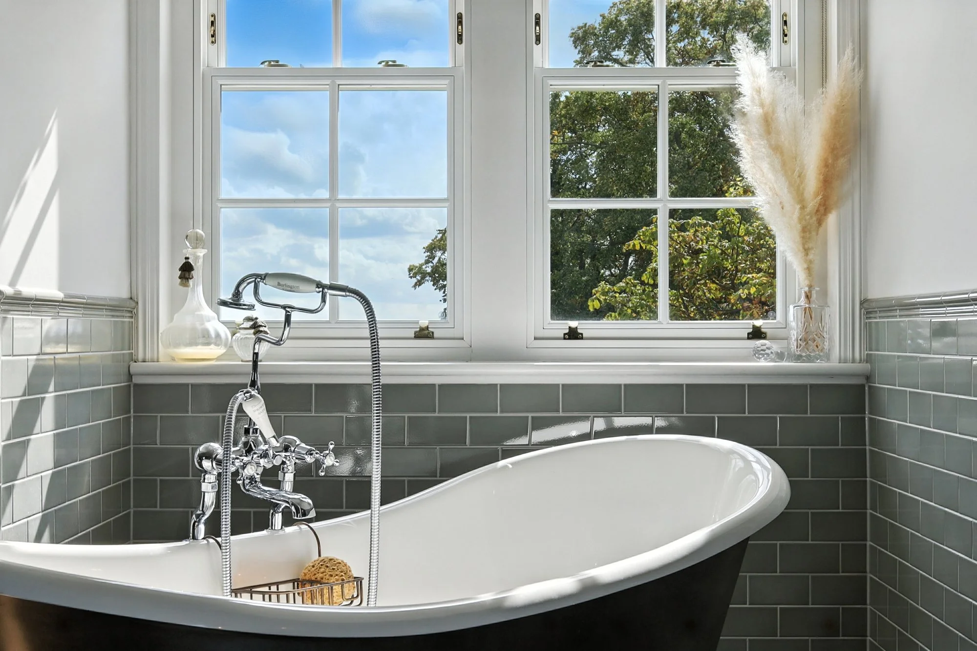 A white clawfoot bathtub with a black exterior in a bathroom, with green tile walls. A window with multiple panes shows blue sky and green trees outside. On the bathtub's edge, there is a wire basket with a sponge, and a vintage-style faucet with a s