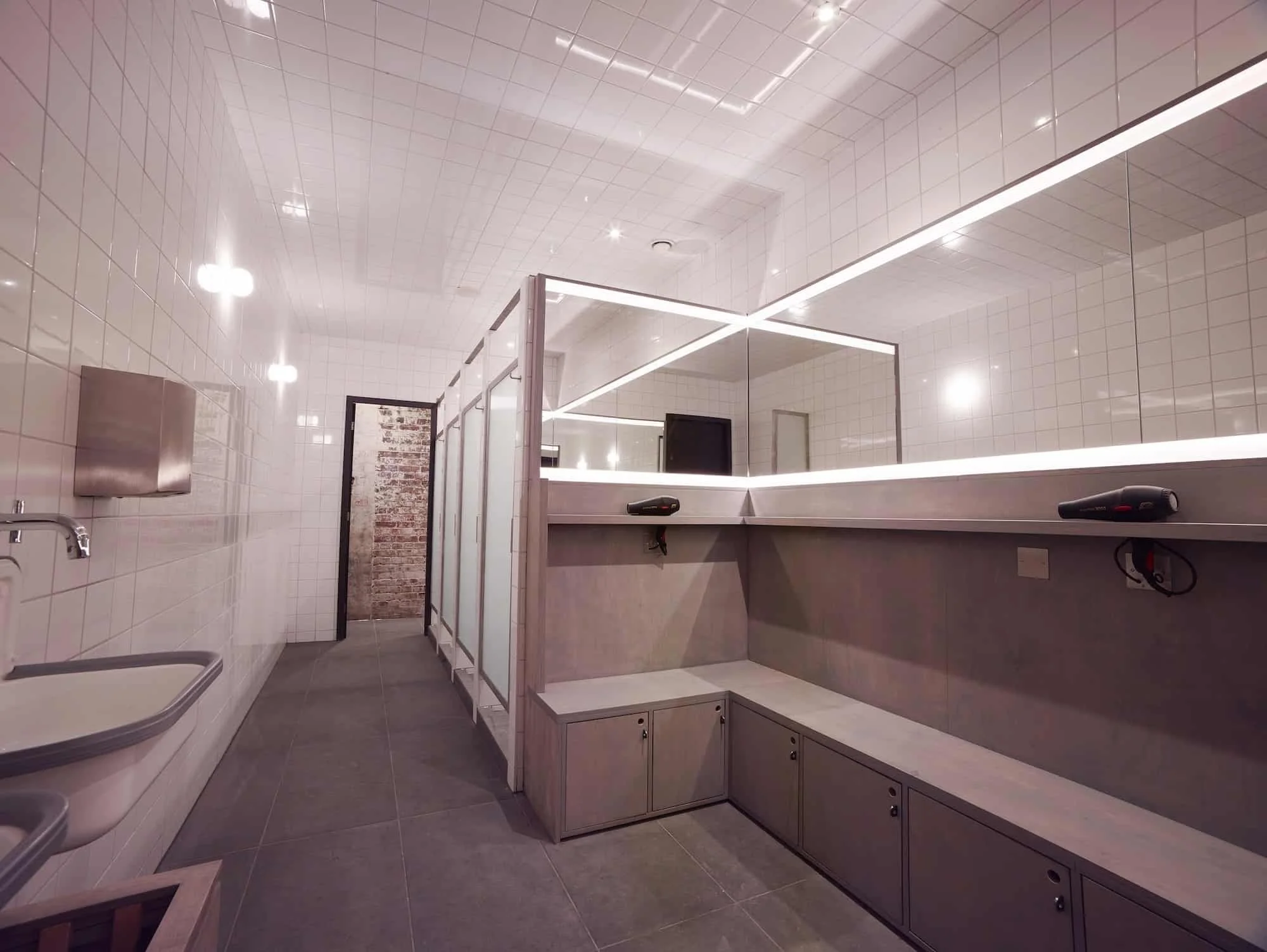 Modern restroom with white tiled walls, gray flooring, a sink, a paper towel dispenser, stalls with frosted glass doors, and a large mirror with light strips.