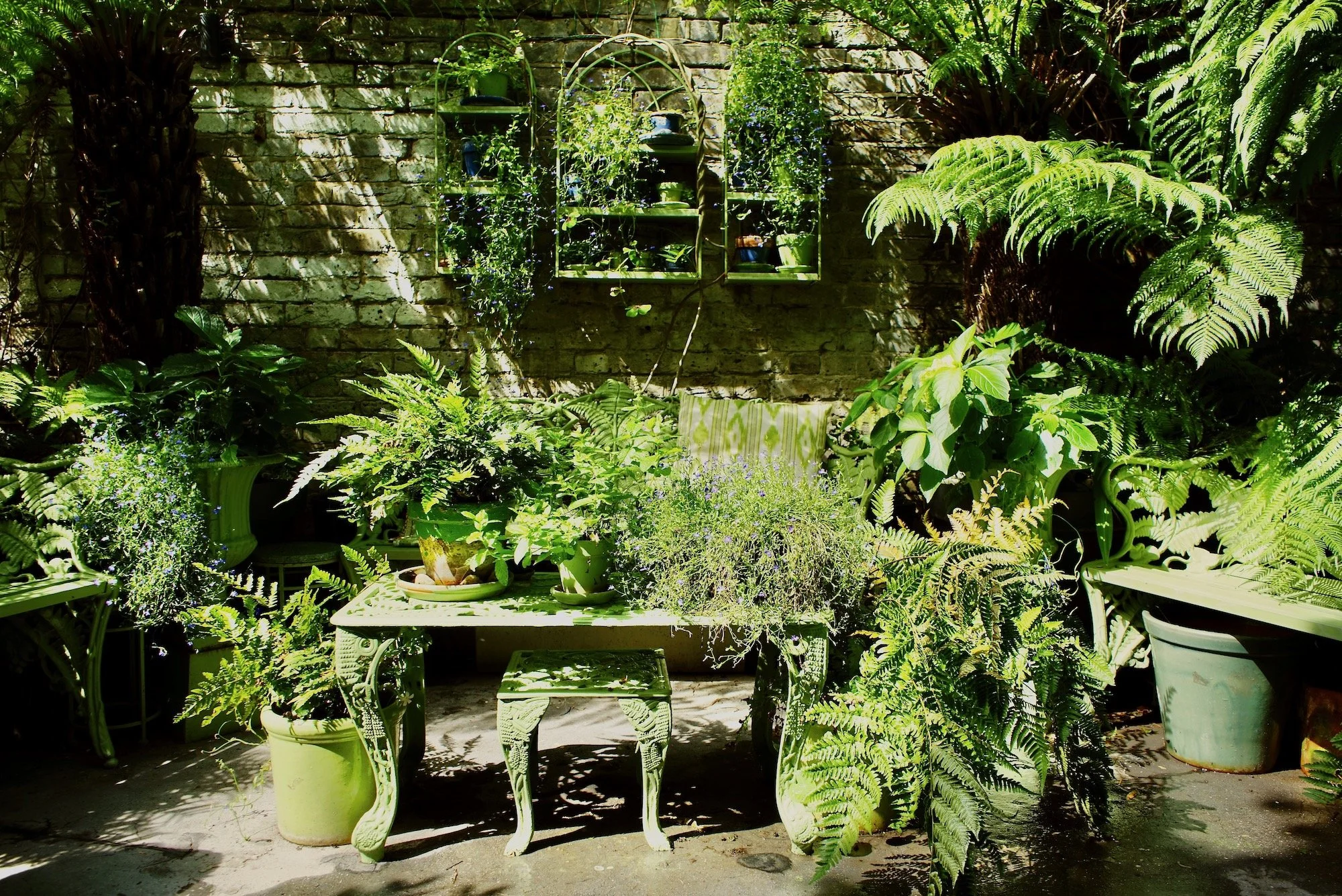 A lush green garden corner with various potted plants, a decorative table, and a brick wall with wall-mounted planters filled with greenery.