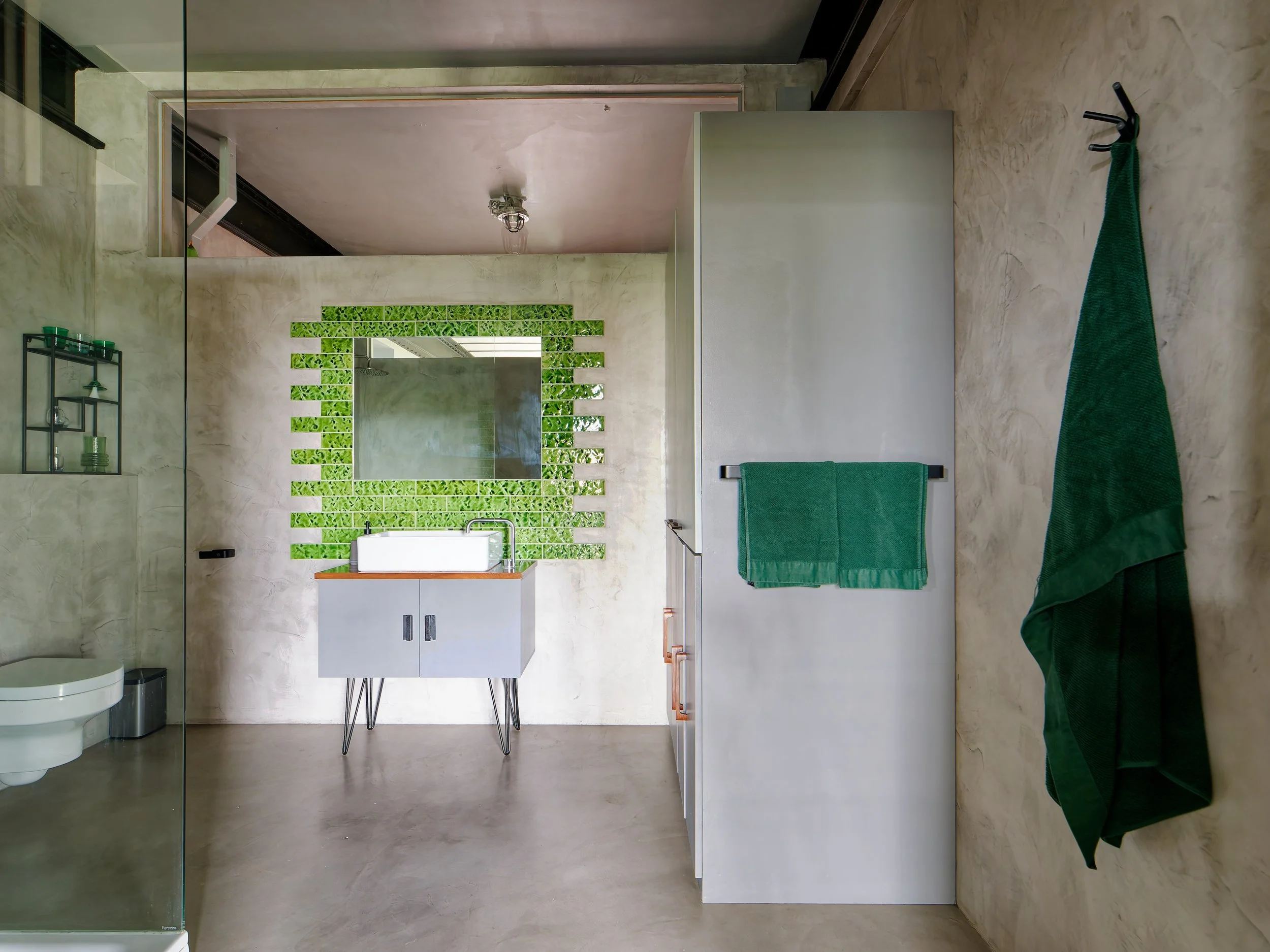 Modern bathroom with a white vanity, green towels, a large mirror, and green wall decor, with a concrete floor and beige textured walls.