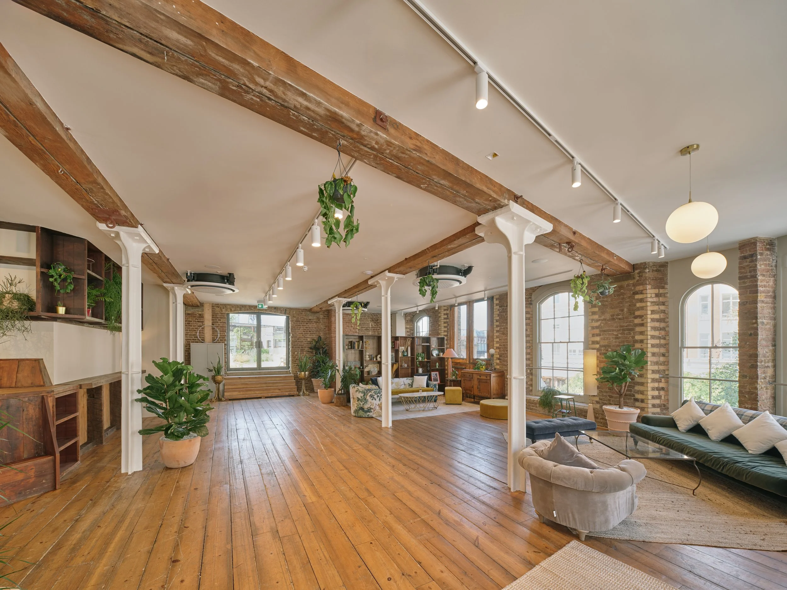 Bright, spacious living room with exposed brick walls, large arched windows, wooden floor, and eclectic furniture. Potted plants and hanging greenery decoratively placed around the room.
