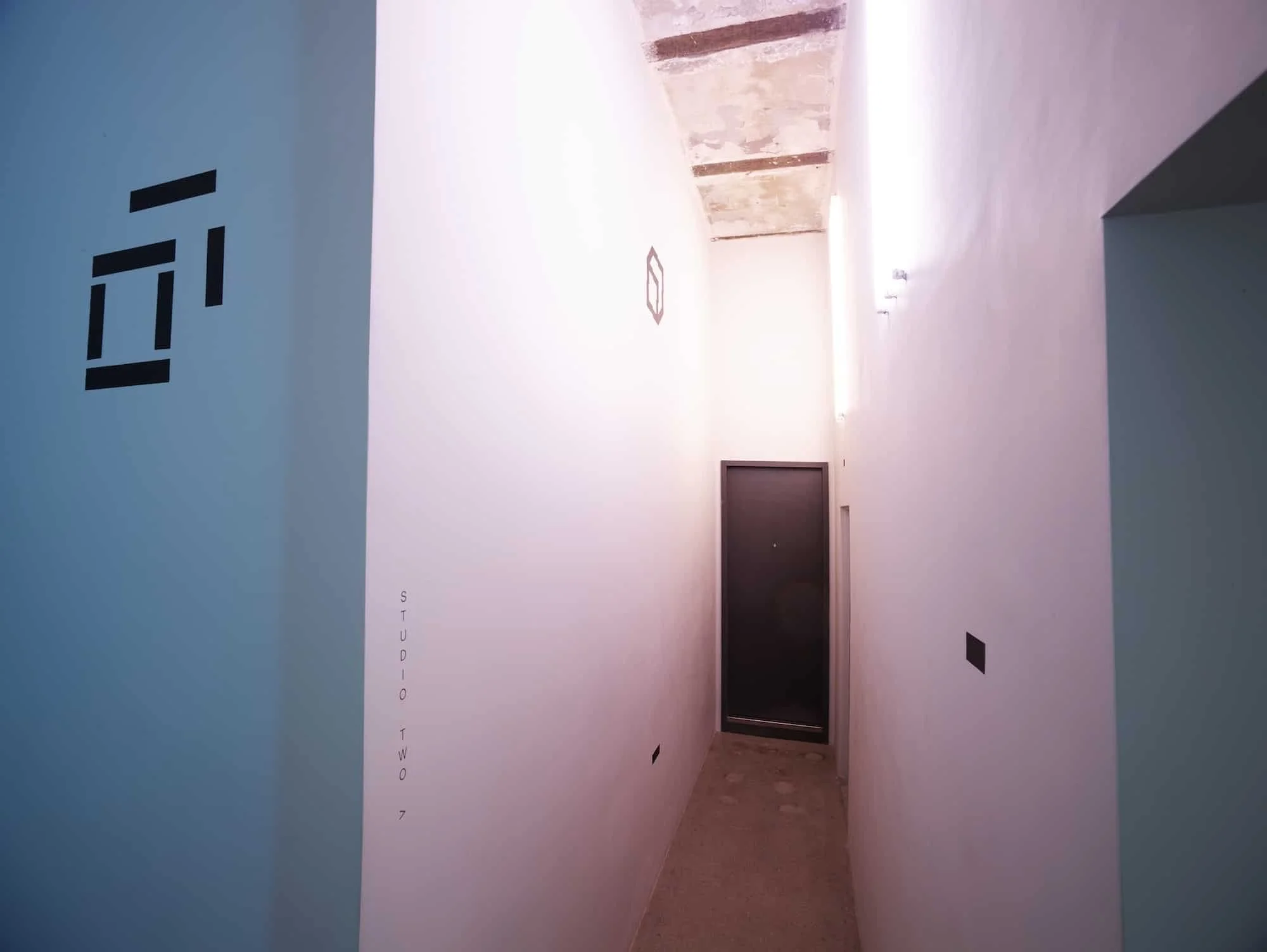 Narrow hallway with white walls and a concrete ceiling, illuminated by a light strip, with black text and designs on the walls, and a dark door at the end.