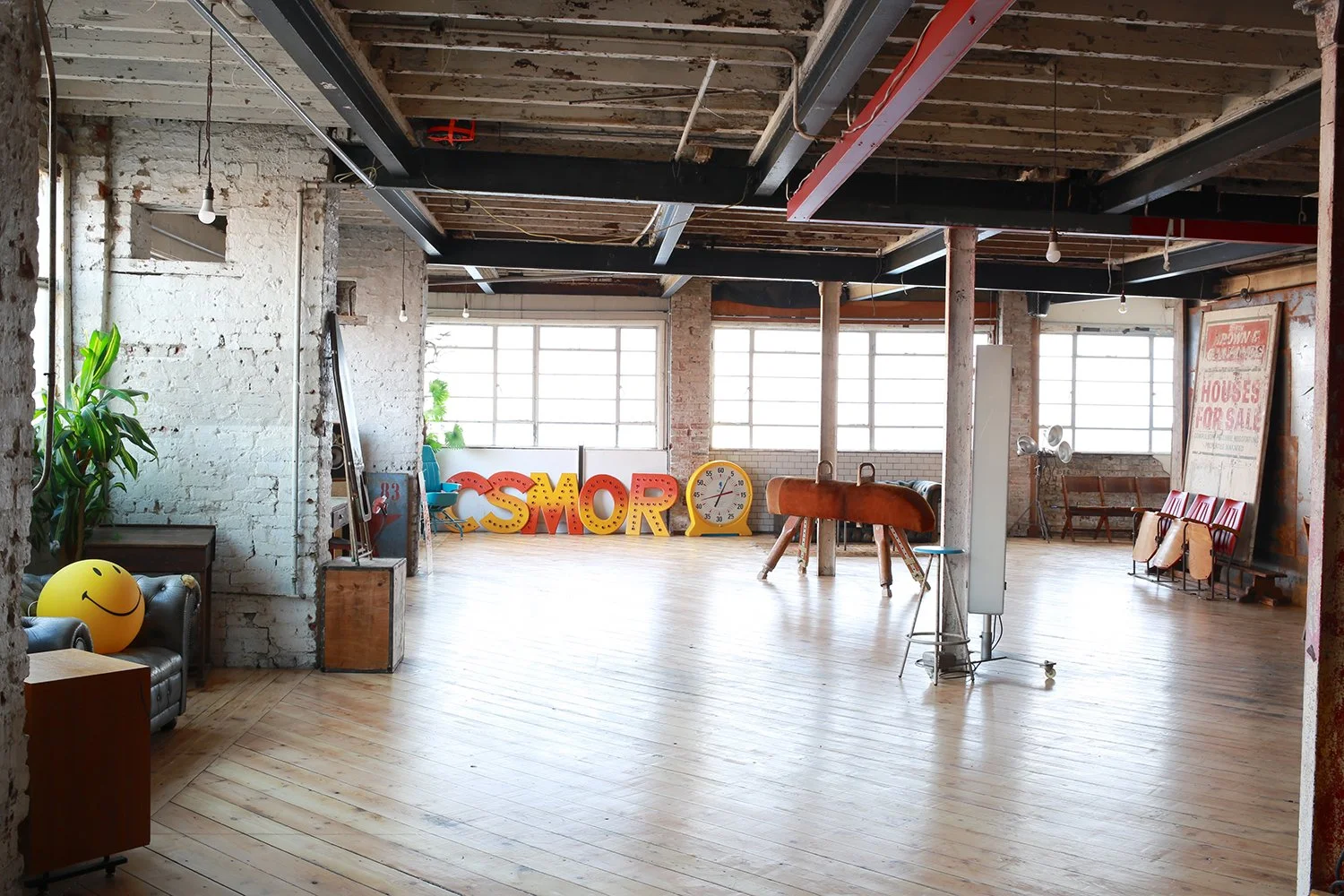 A spacious, industrial-style room with exposed brick walls, large windows, wooden floors, and various decorative items including colorful letters spelling 'SMOR', a large smiley face ball, and vintage furniture.