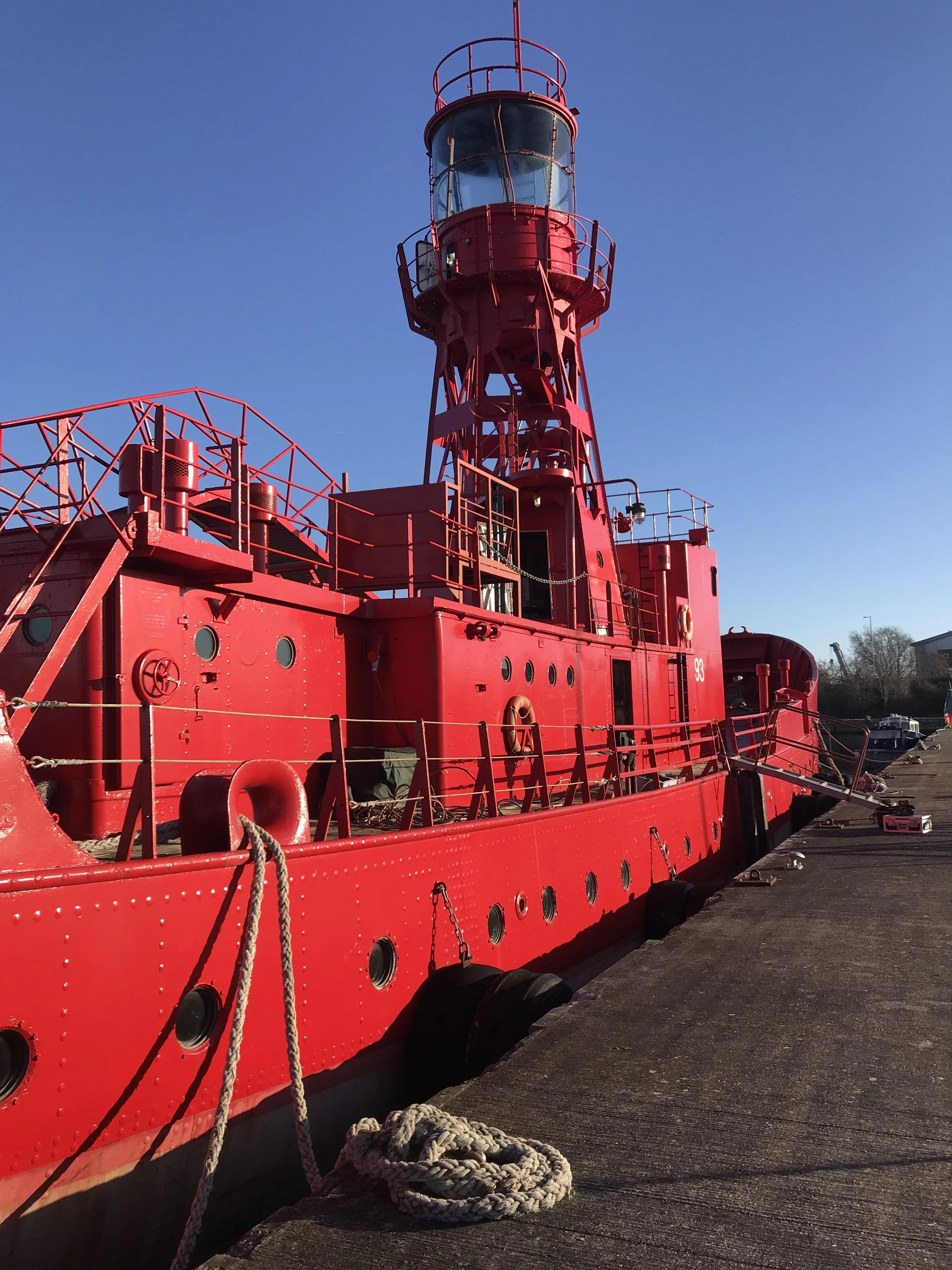 A bright red lighthouse ship moored along the dockside, with a tall lighthouse tower structure at the stern, under a clear blue sky.