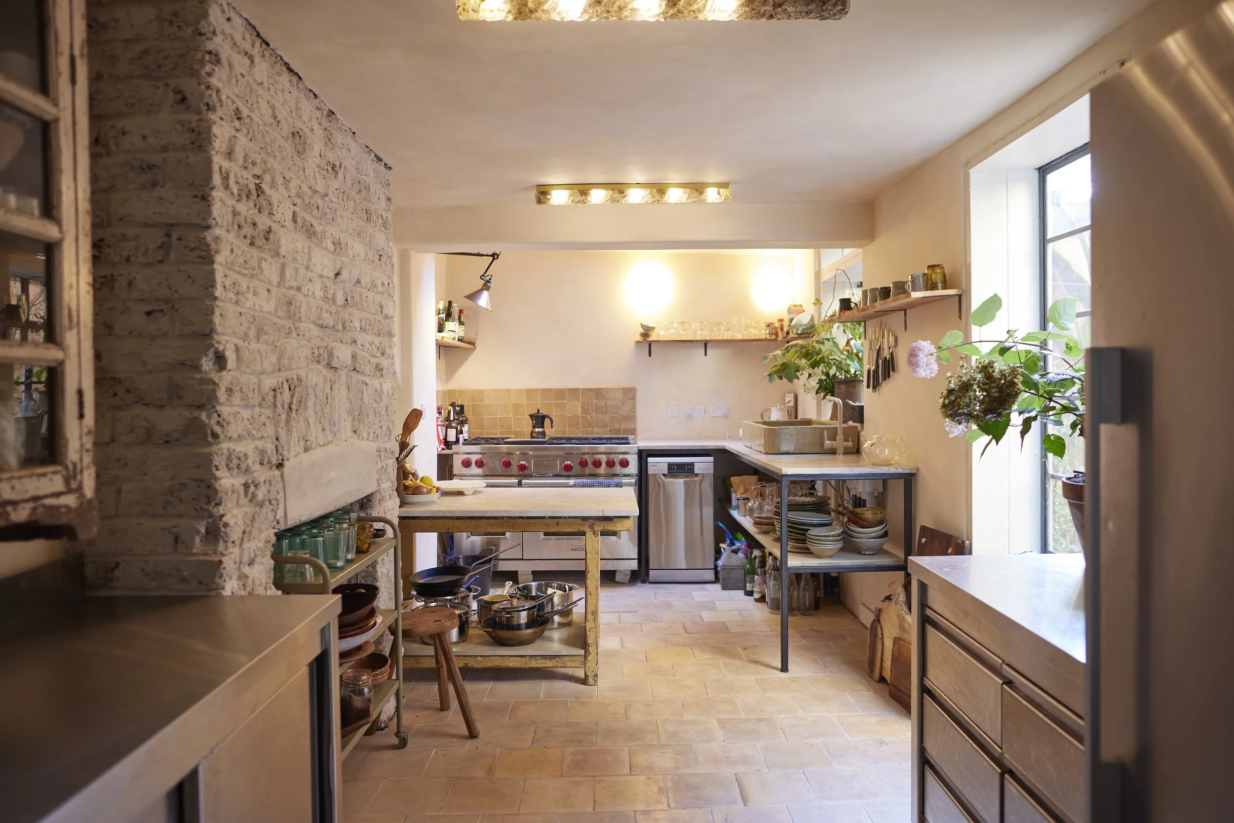 A cozy kitchen with a rustic white brick wall on the left, open shelving with plants and jars, a stainless steel stove with red knobs, a kitchen sink, and various pots and dishes. Large window on the right lets in natural light, and the space has war