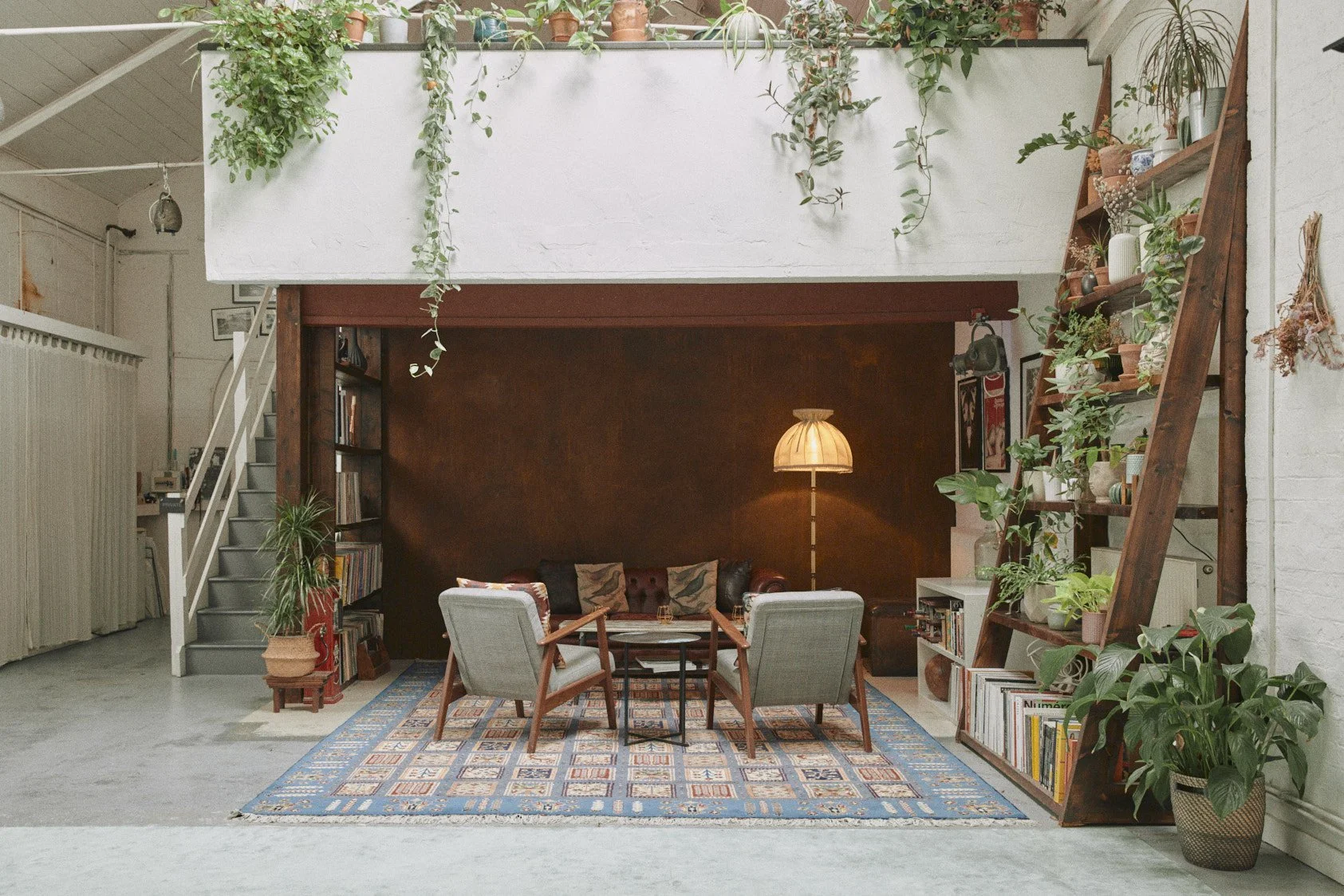 Living room with a dark brown accent wall, a vintage lamp, two green armchairs, a brown leather sofa, a patterned area rug, and shelves filled with books and plants.