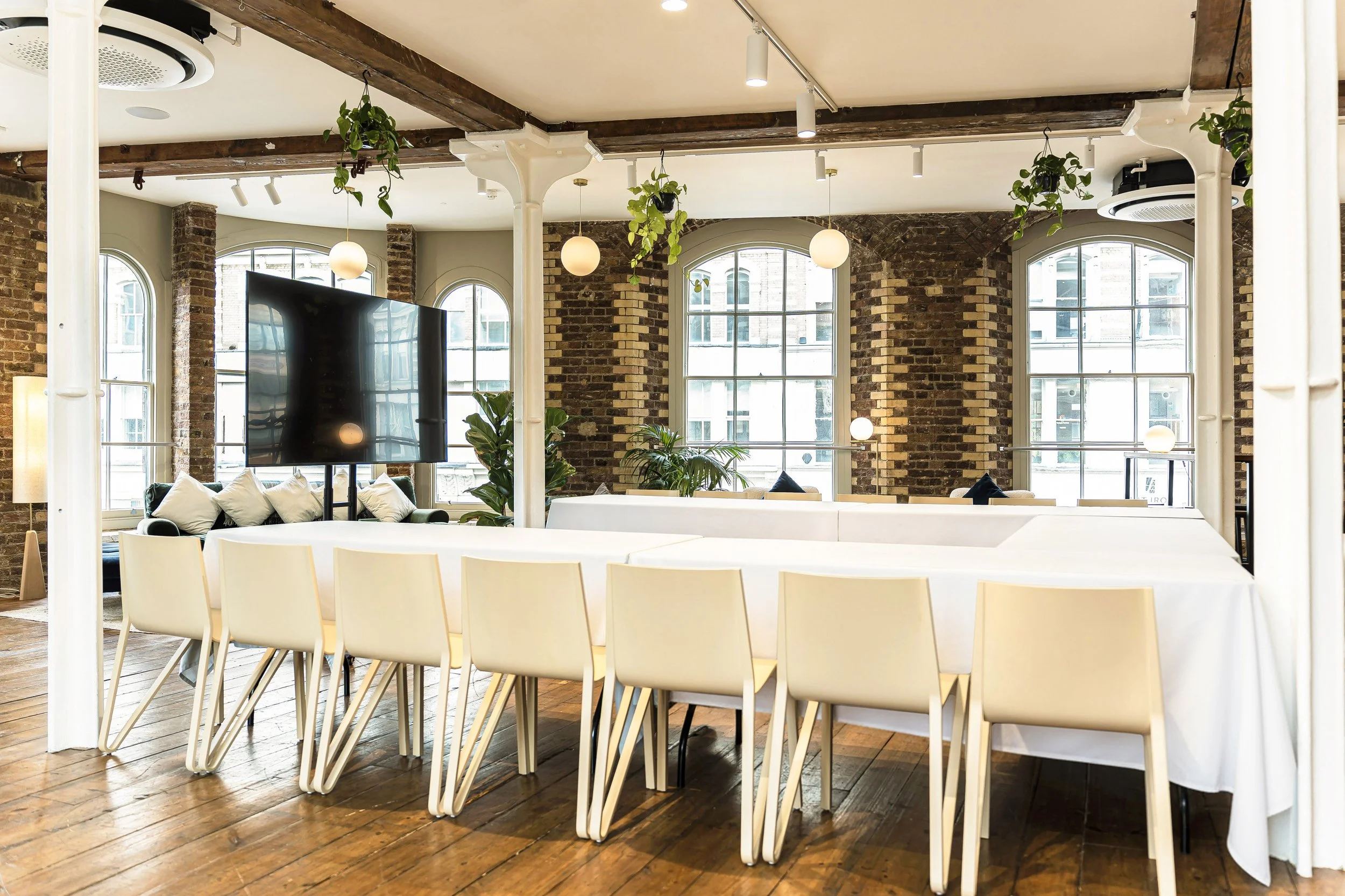 Interior of a modern event space or conference room with a long table covered with a white tablecloth, surrounded by beige chairs with a wooden floor, large windows with brick walls, hanging plants, and a large flat-screen TV.