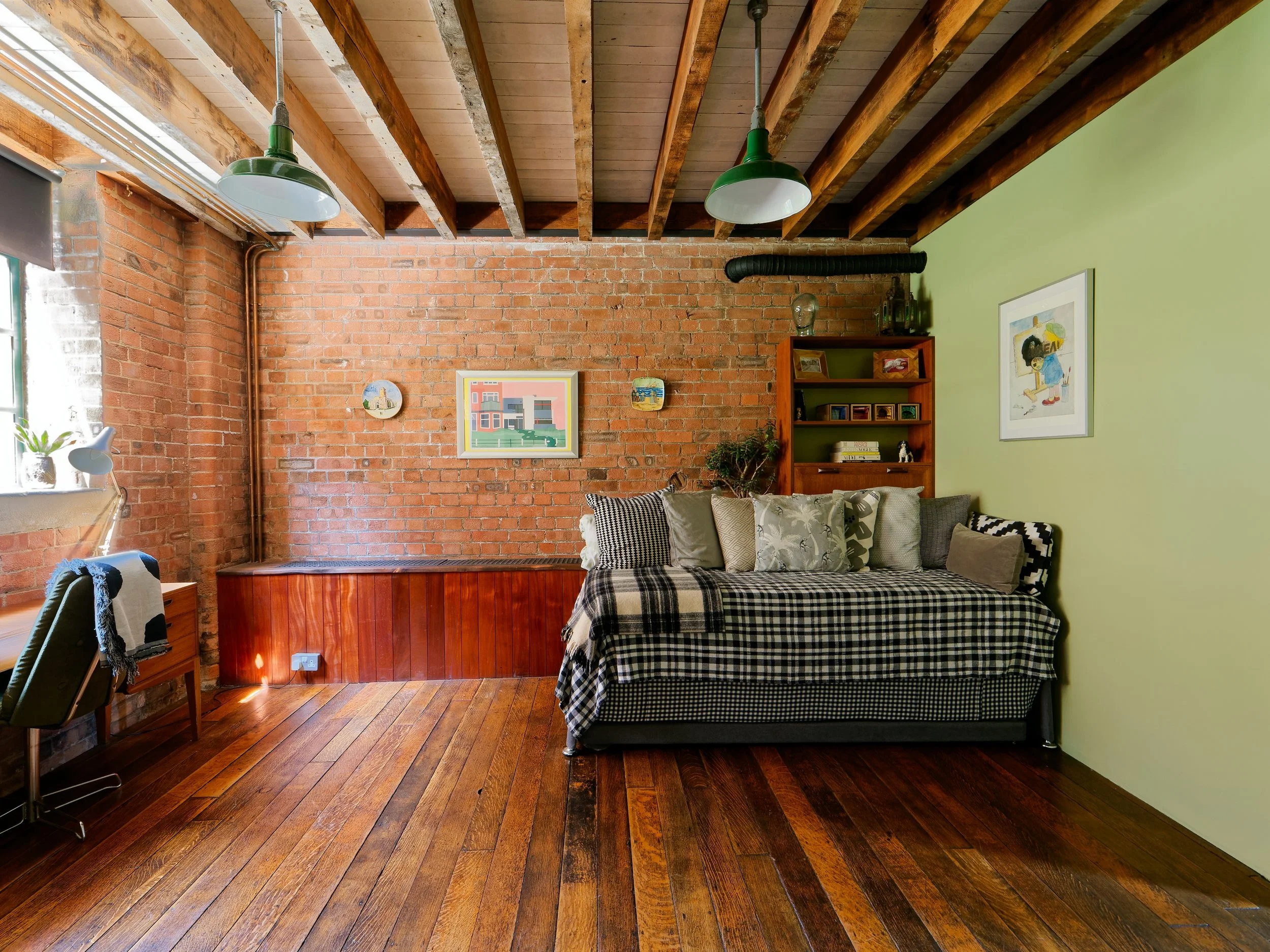 Living room with exposed brick and painted walls, hardwood floor, ceiling with wooden beams, and mixed furniture including a daybed with patterned pillows, a green wall, and a small bookshelf.