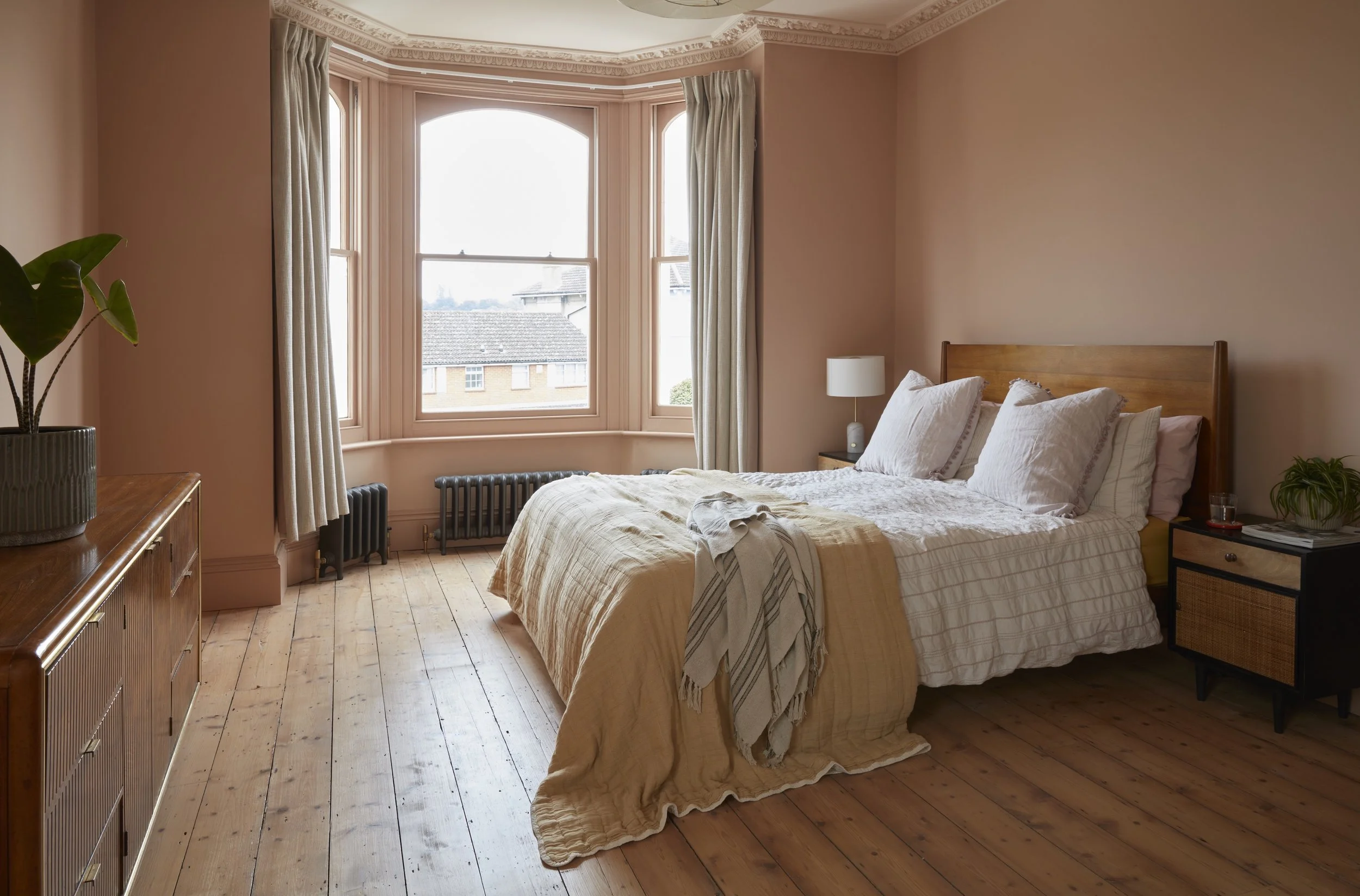A cozy bedroom with a large wooden bed, white bedding, and a beige blanket. There is a white nightstand with a lamp, a plant, and a glass of water. The room has wooden floors, pink walls, and a big bay window with cream curtains, overlooking neighbor