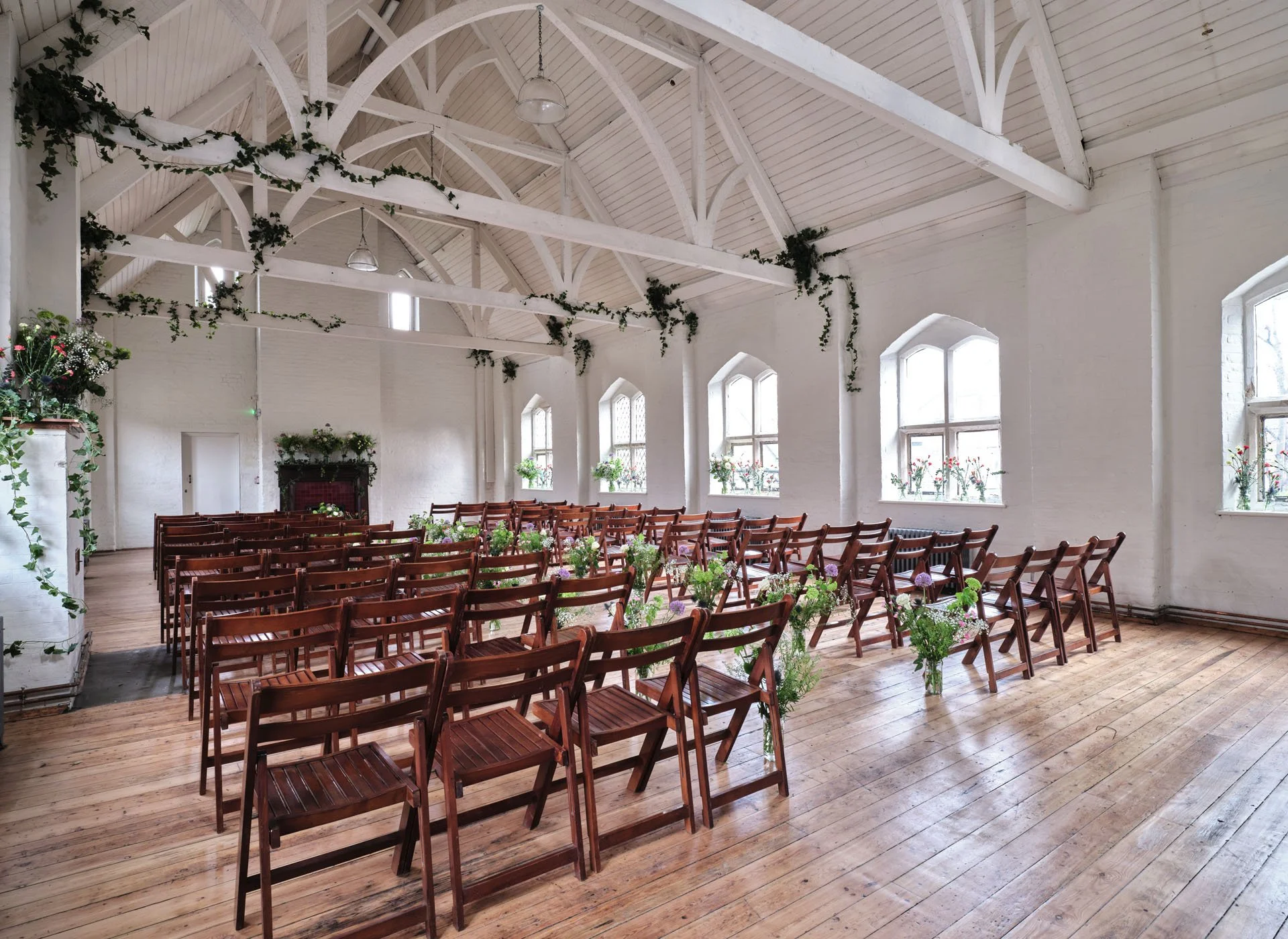 An empty room prepared for a wedding or ceremony, with wooden chairs arranged in rows, decorated with potted flowers, and greenery hanging from the white ceiling beams.