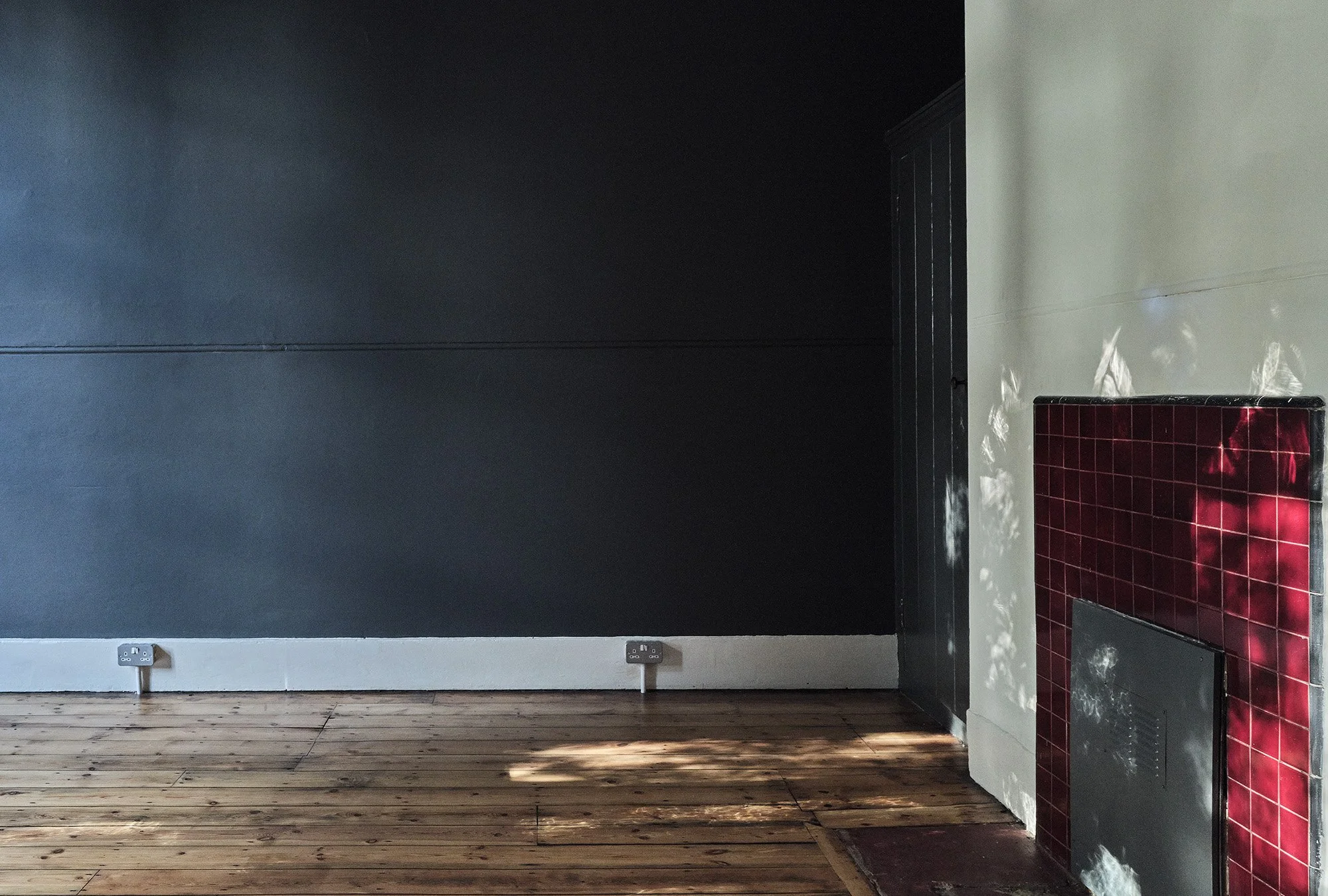 Interior of a room with a wood floor, a black wall, and a fireplace with red tiles, with a door and some electrical outlets visible.
