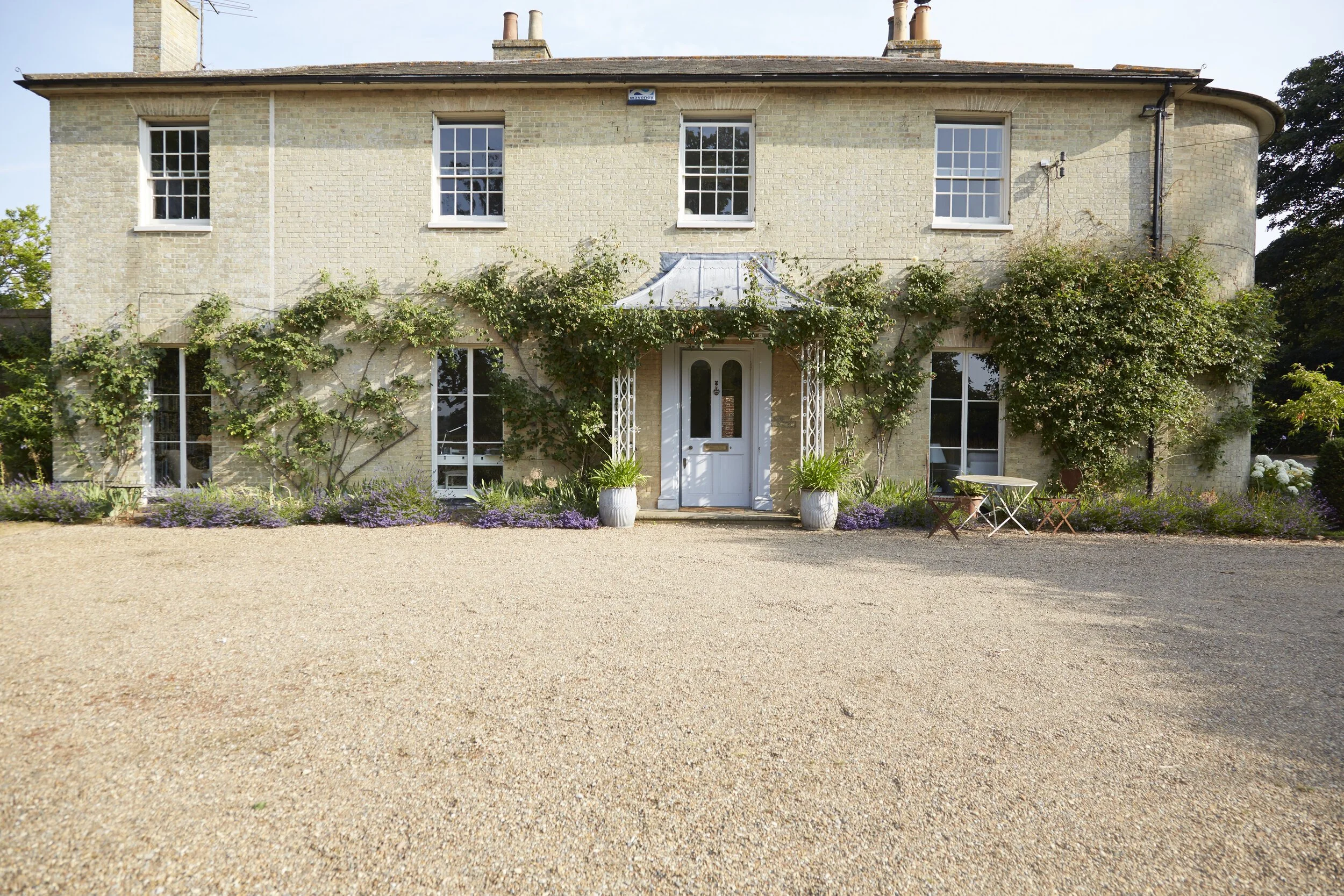 A two-story brick house with a white front door, surrounded by green ivy and purple flowers. There is a gravel driveway in front and outdoor furniture to the right. The house has multiple windows and chimney stacks on the roof.