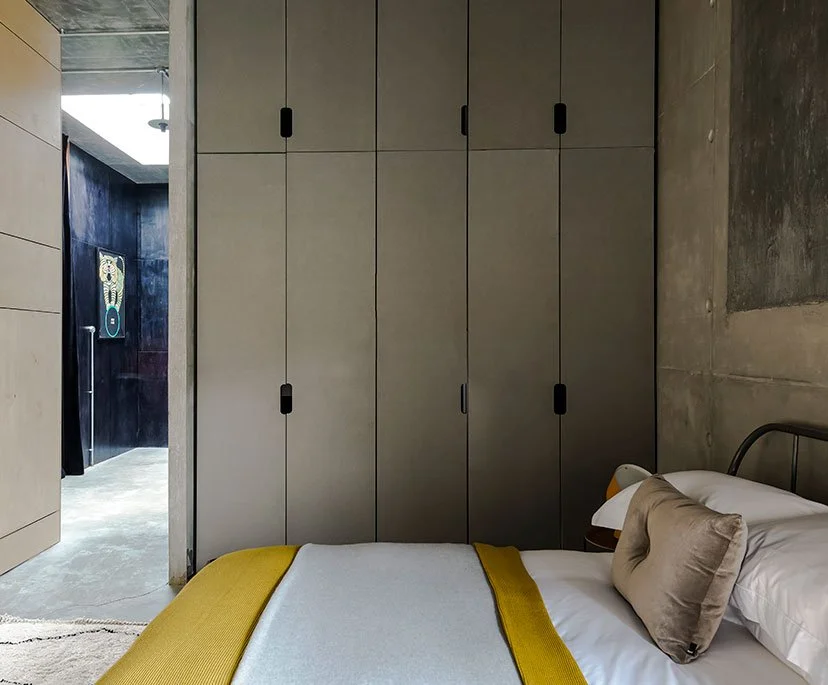 A bedroom with a bed, beige pillows, and a yellow blanket, next to a grey wardrobe, and a doorway showing a part of an adjacent room with a black wall and a piece of modern artwork.