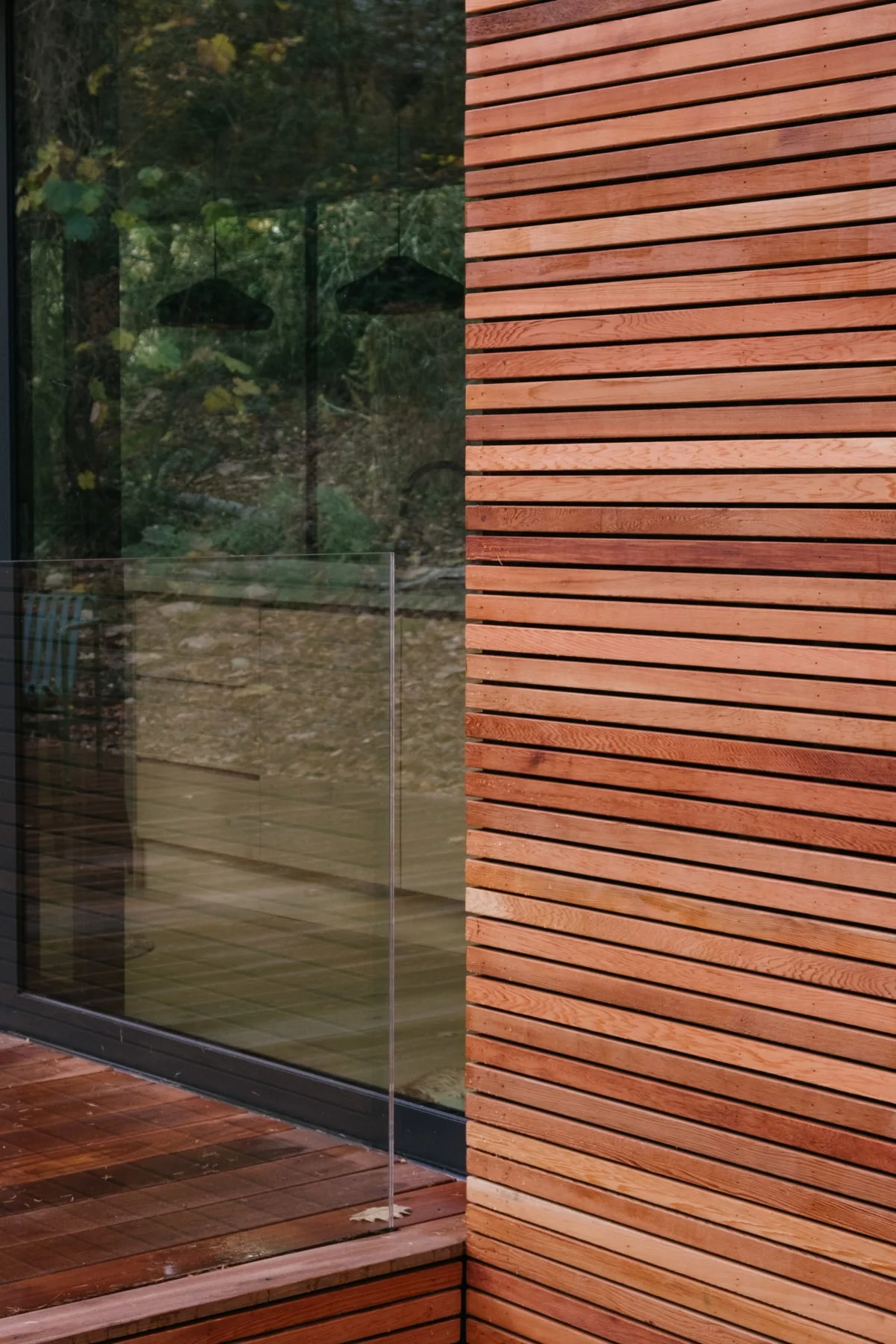 Close-up of a modern building corner with horizontal wooden slat exterior wall, large glass window reflecting trees and outdoor furniture, and wooden flooring.