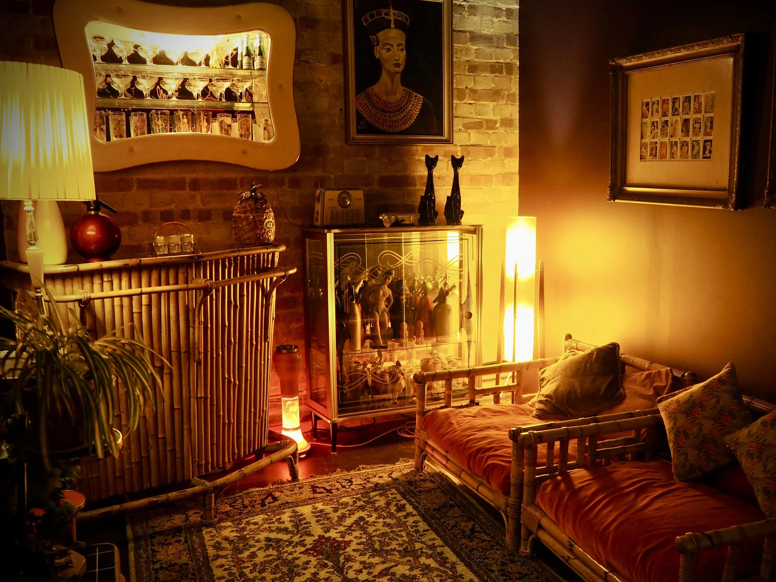 A cozy living room with warm yellow lighting, including a bamboo sofa with pillows, a patterned area rug, a glass display cabinet with figurines, and a brick wall decorated with artwork and framed pictures.