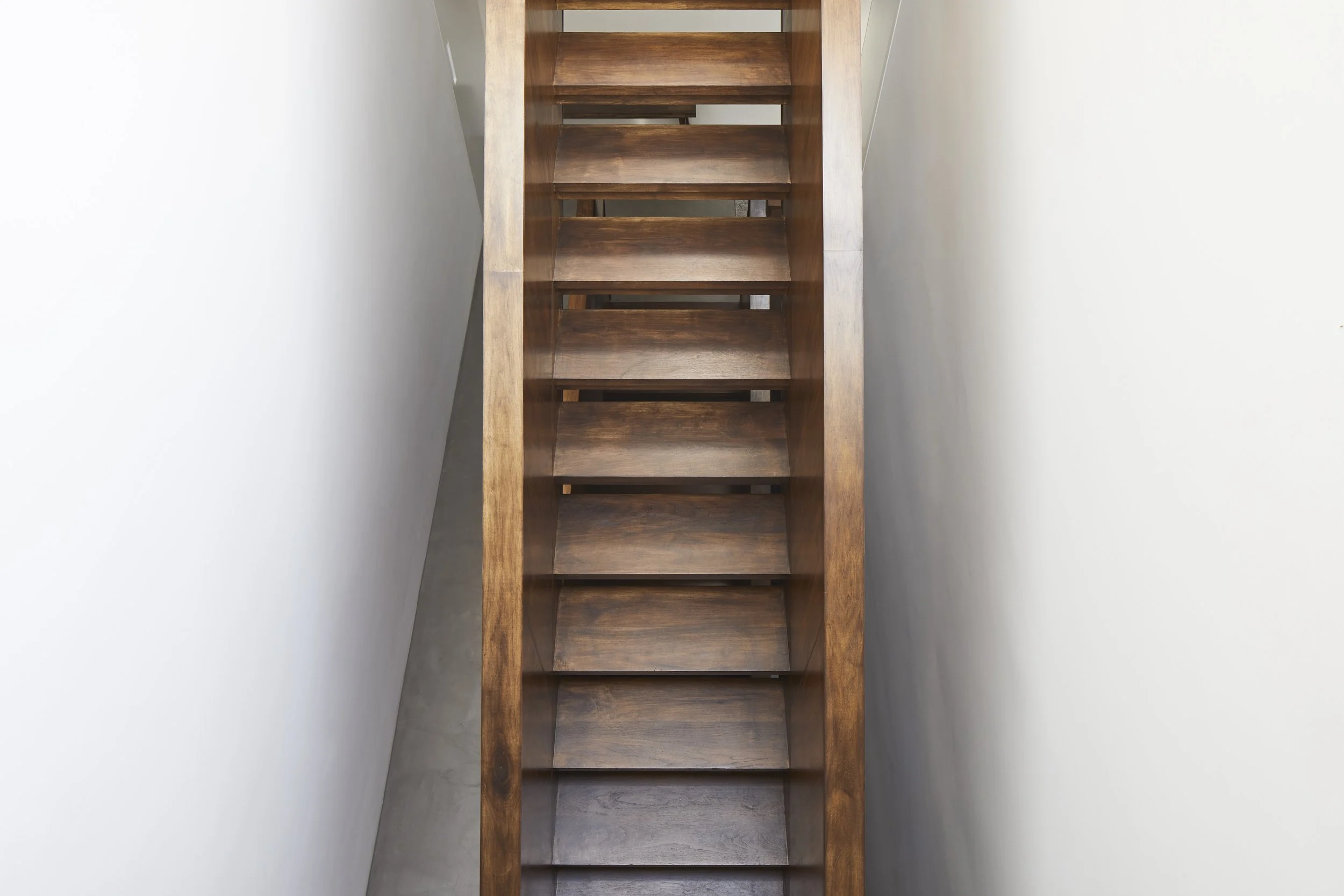 A top-down view of a wooden staircase with open risers, positioned between white walls.