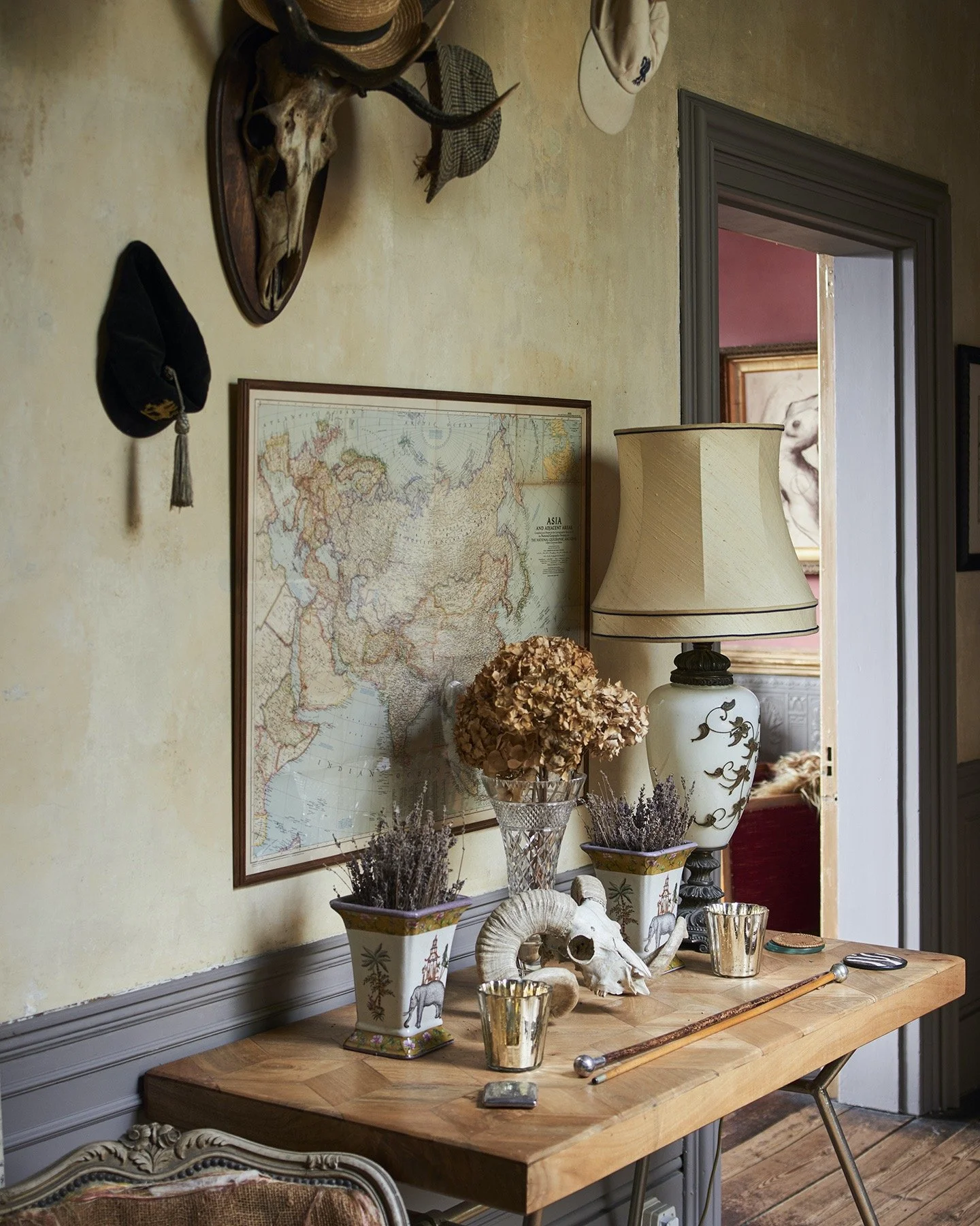 A wooden table decorated with potted lavender, dried flowers, a ram skull, and ceramic vases, with a world map on the wall, a lamp, and hats hanging above.