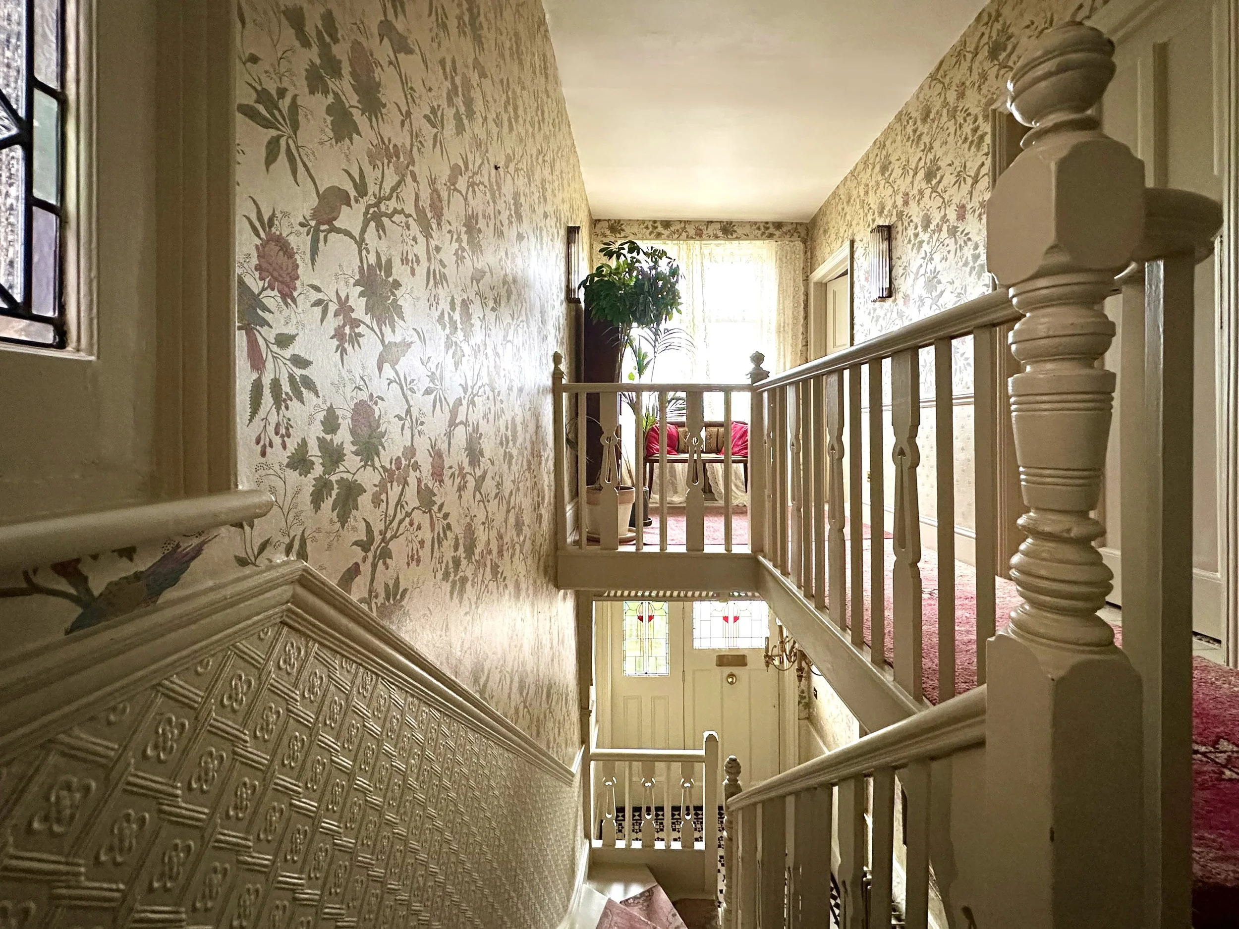A view from the stairway landing looking into a hallway on the upper floor. The hallway has floral wallpaper, a window with curtains, and a small sitting area with a red sofa and a potted plant. The staircase and railing are painted white.