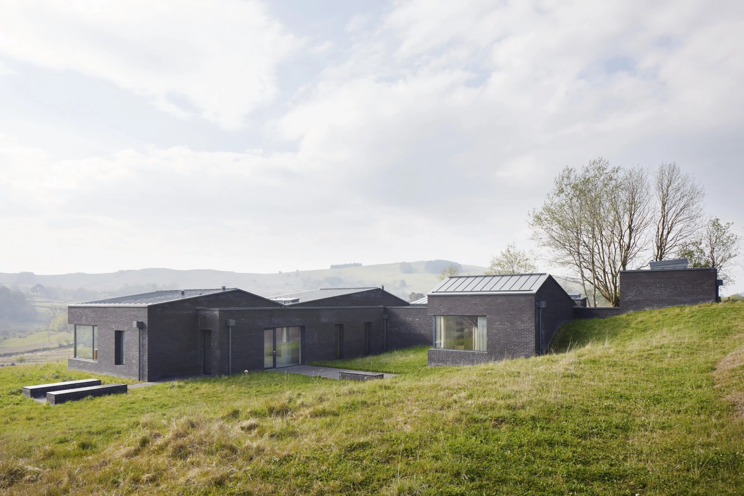Modern black brick house with large windows situated on a grassy hillside with trees in the background and hills in the distance under a partly cloudy sky.
