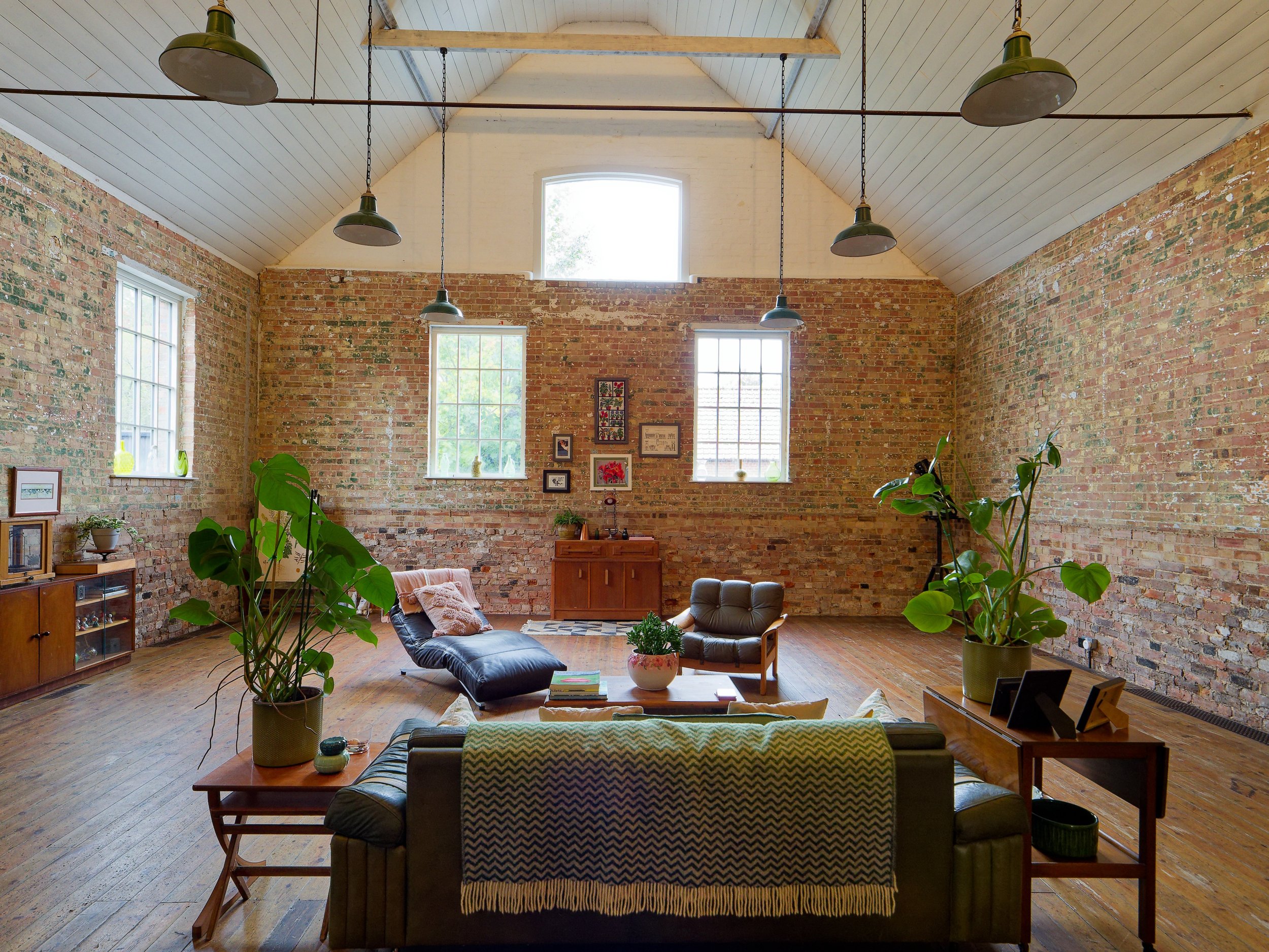 A spacious living room with exposed brick walls, large windows, hardwood floors, and a vaulted ceiling. It has a mix of mid-century modern furniture including a black leather lounge chair, a cushioned armchair, a long sofa covered with a throw, and s