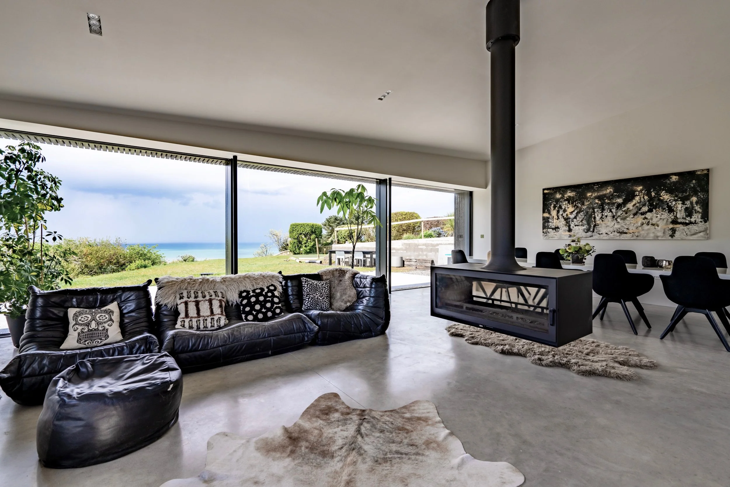 Living room with large window showing green lawn and ocean outside. Black leather sofa with patterned and furry pillows, cowhide rug, and modern fireplace. Dining area with black chairs and art on the wall.