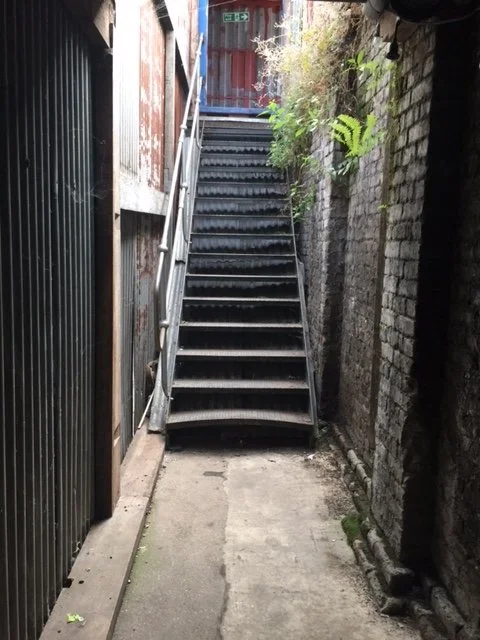 Narrow outdoor stairway with metal railings, between brick and wooden walls, leading up to a gate. Some greenery visible on the right side.