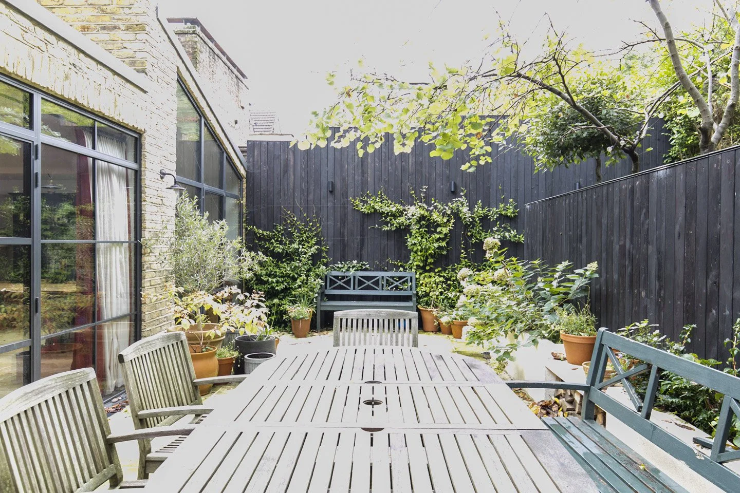 Empty outdoor patio with wooden dining table, multiple garden benches, and potted plants along a black wooden fence, surrounded by greenery and trees.
