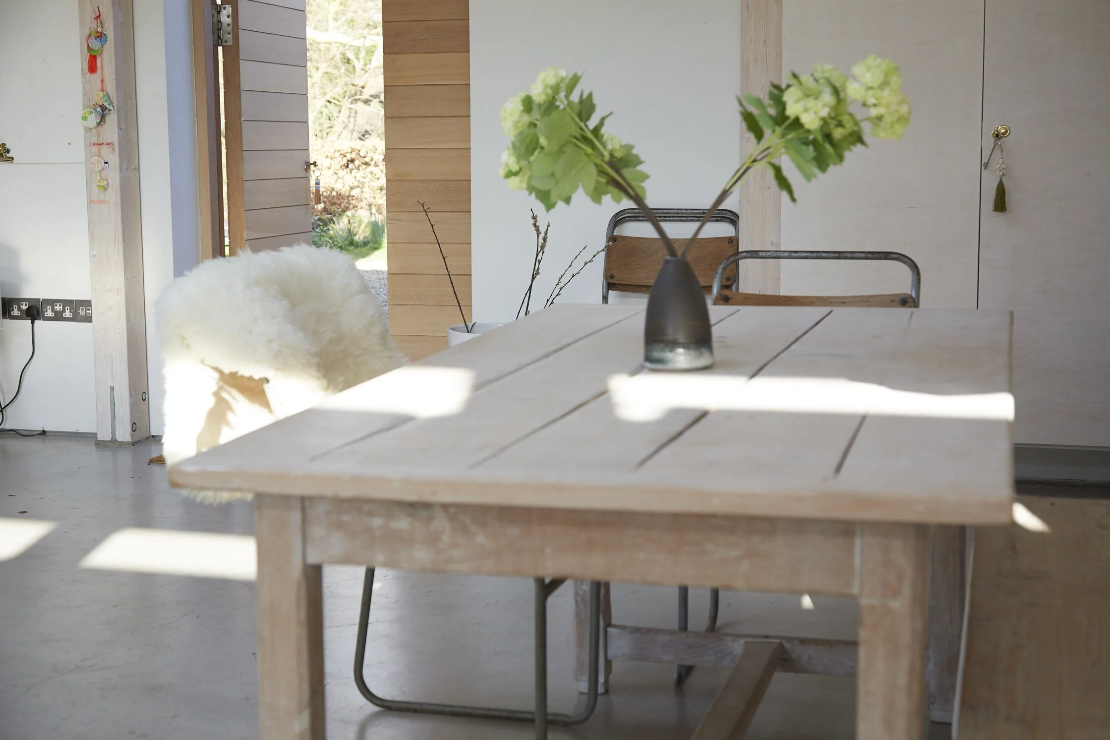 A wooden dining table with a black vase holding green hydrangea flowers, a white Sheepskin rug on a chair, and two black chairs behind the table in a bright, modern interior with natural light.