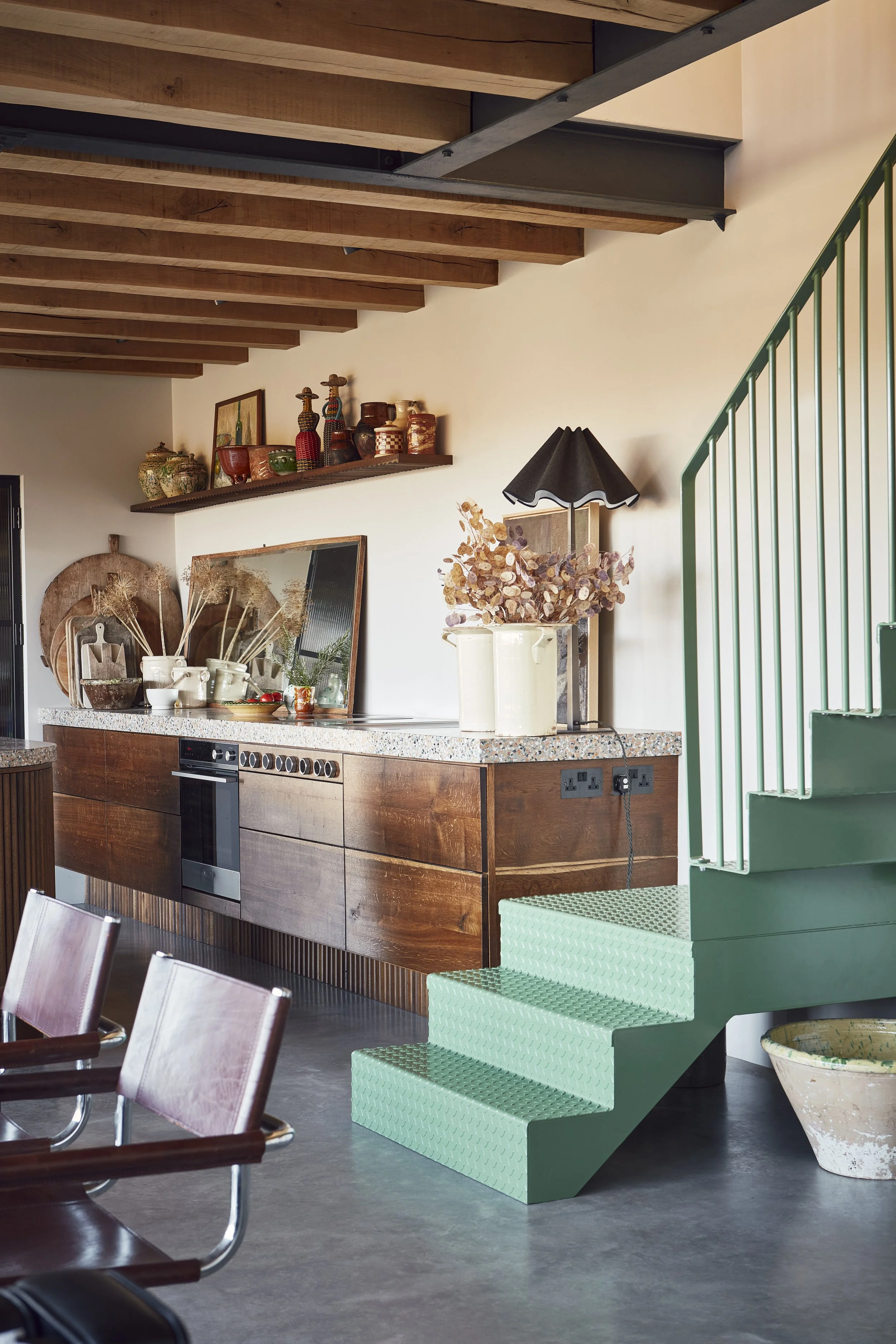 Interior of a modern kitchen with wooden ceiling beams, a countertop with various decorative pots, vases, and a mirror, and a green staircase leading upstairs.