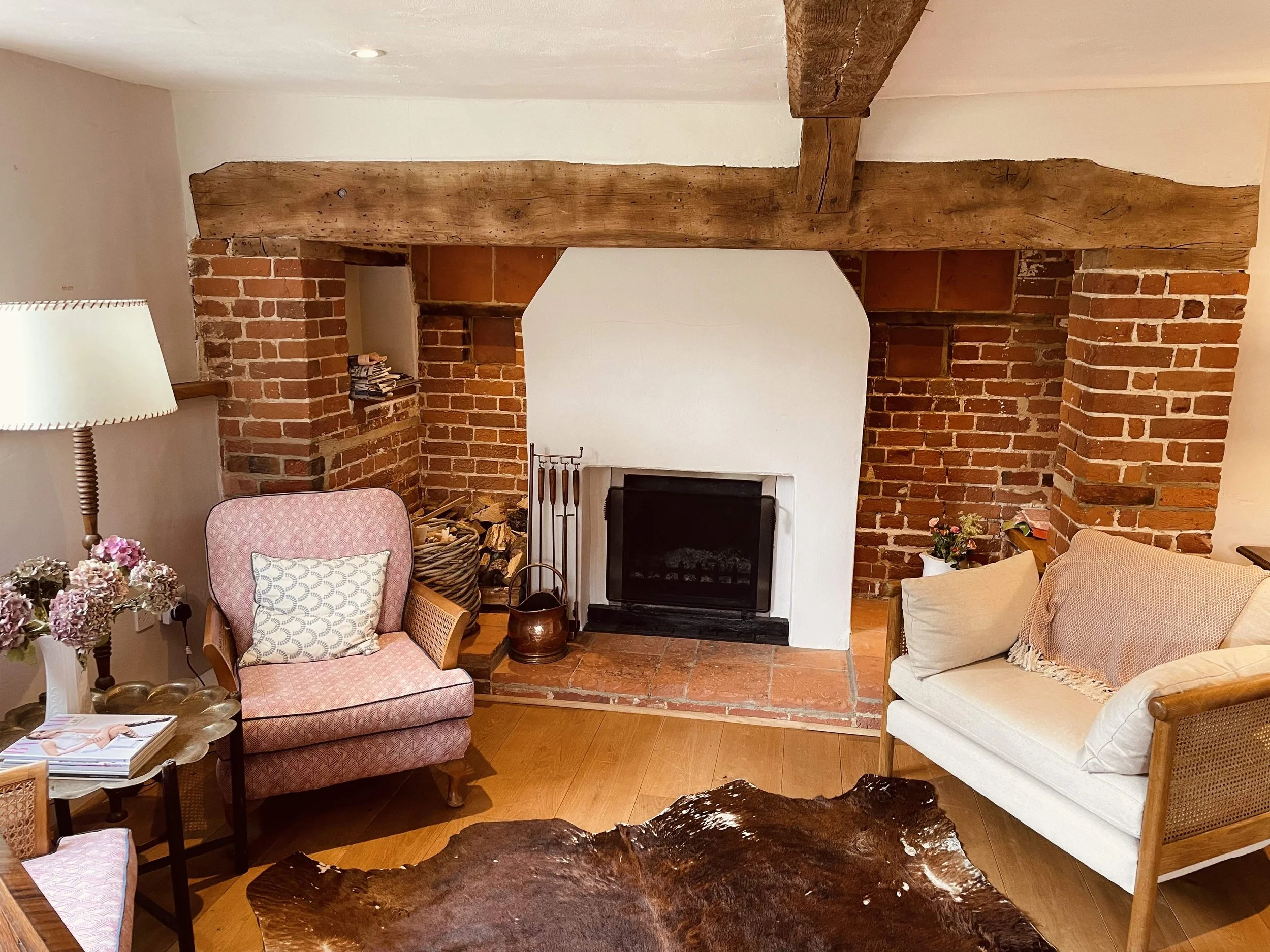 Living room with exposed brick and wooden beams above a fireplace. Furniture includes pink floral patterned armchair, white sofa, and wooden chairs. Decor features flowers, pillows, and books.