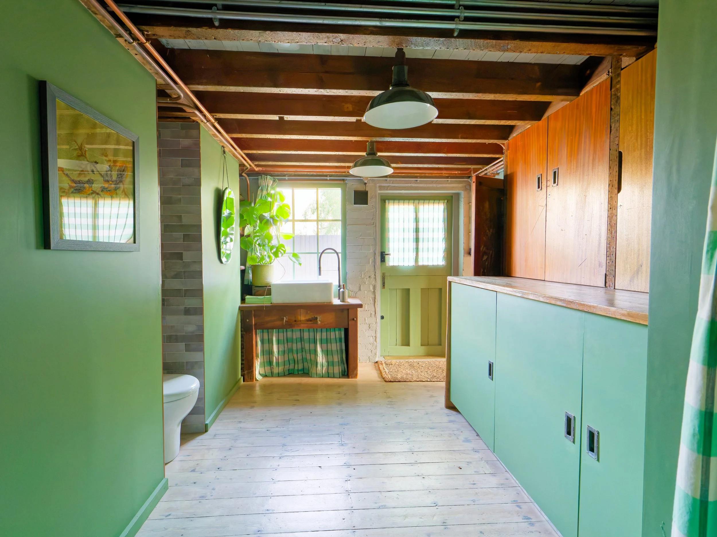 Bright, rustic kitchen with light green walls, wooden ceiling beams, and large windows. Features a white farmhouse sink, potted plant, and wooden and green storage cabinets, with natural light streaming in.