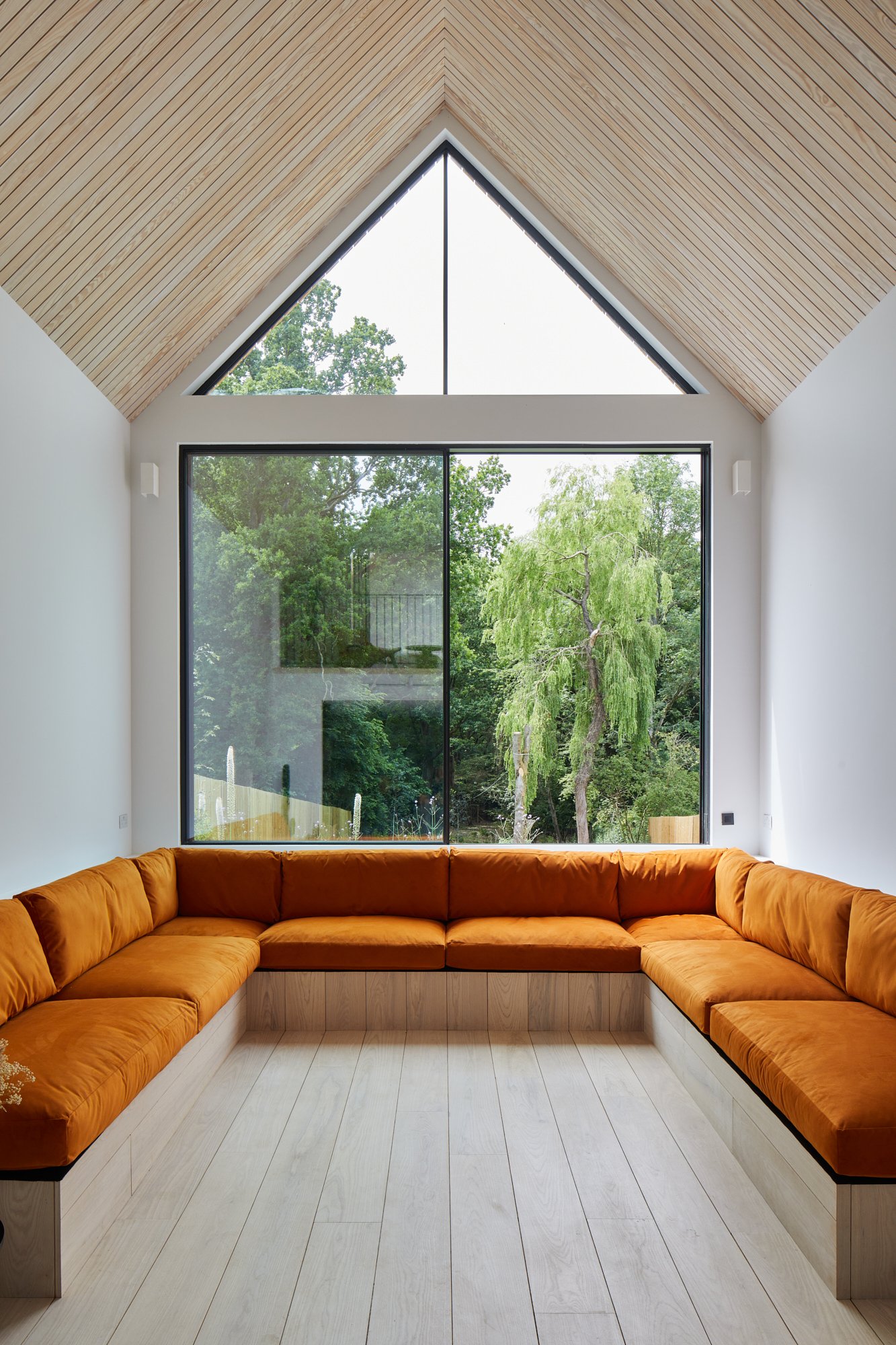 A modern living room with a large L-shaped orange velvet sofa, white walls, and a high vaulted ceiling with wooden paneling. The room features big windows looking out onto a lush green landscape.