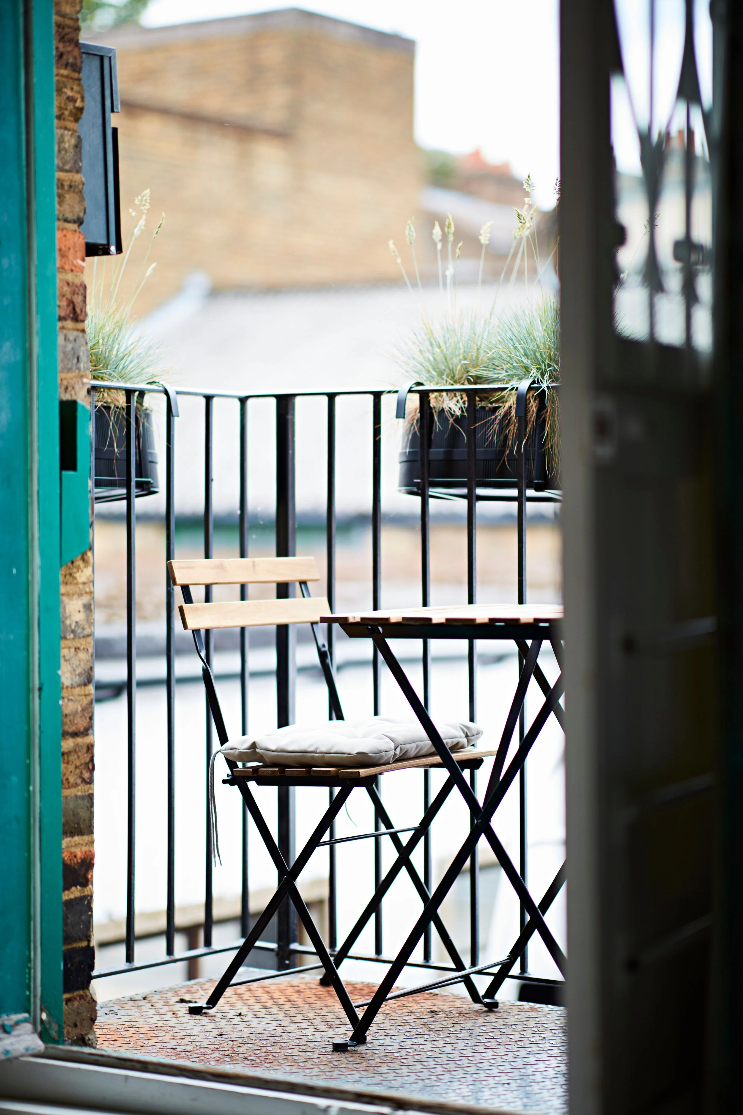View of a small balcony with two potted plants, a wooden and metal chair with a cushion, and a small table, visible from inside a building.