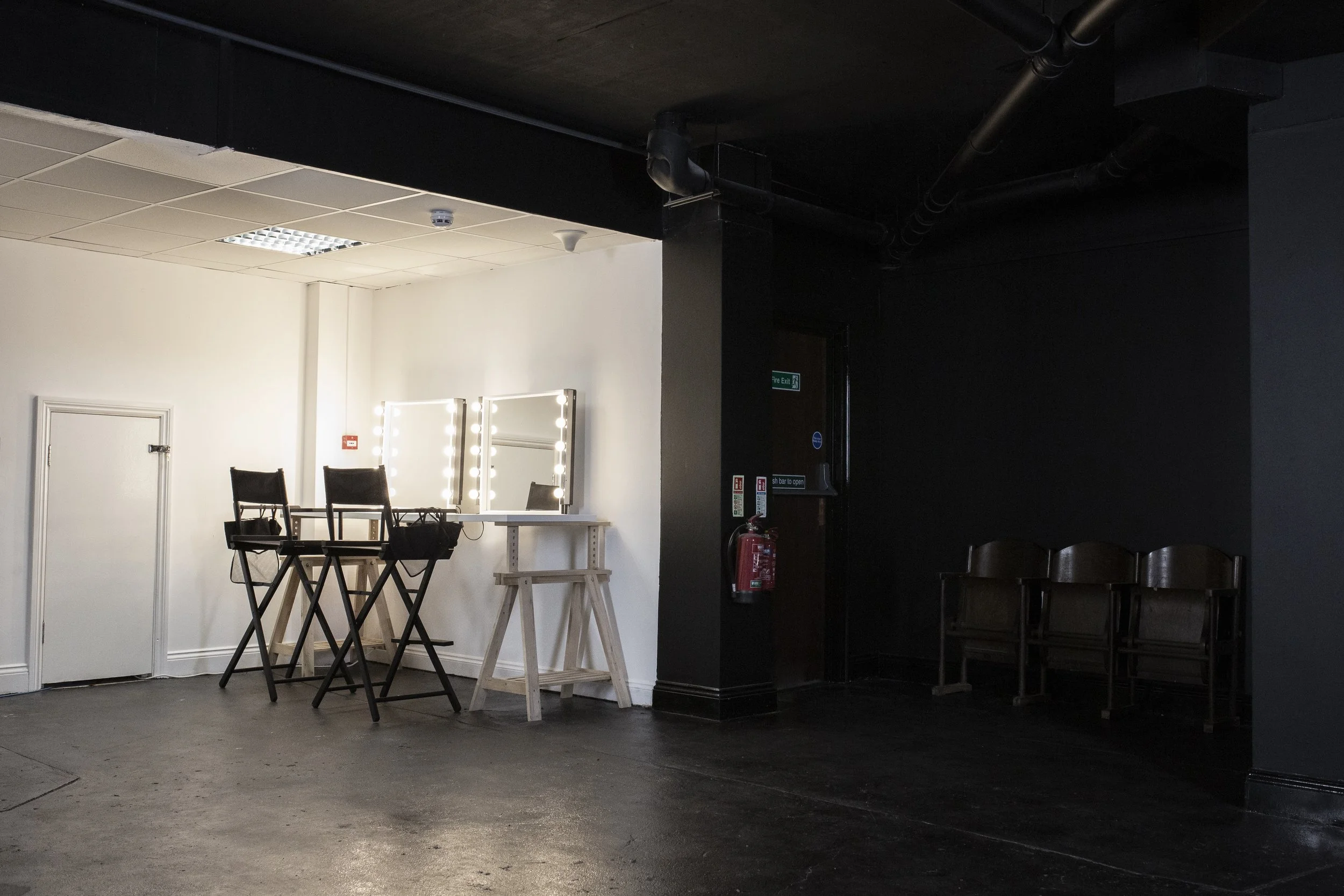 An interior space divided into a well-lit white side with makeup mirrors and chairs, and a dark side with empty chairs and fire safety and exit signs.