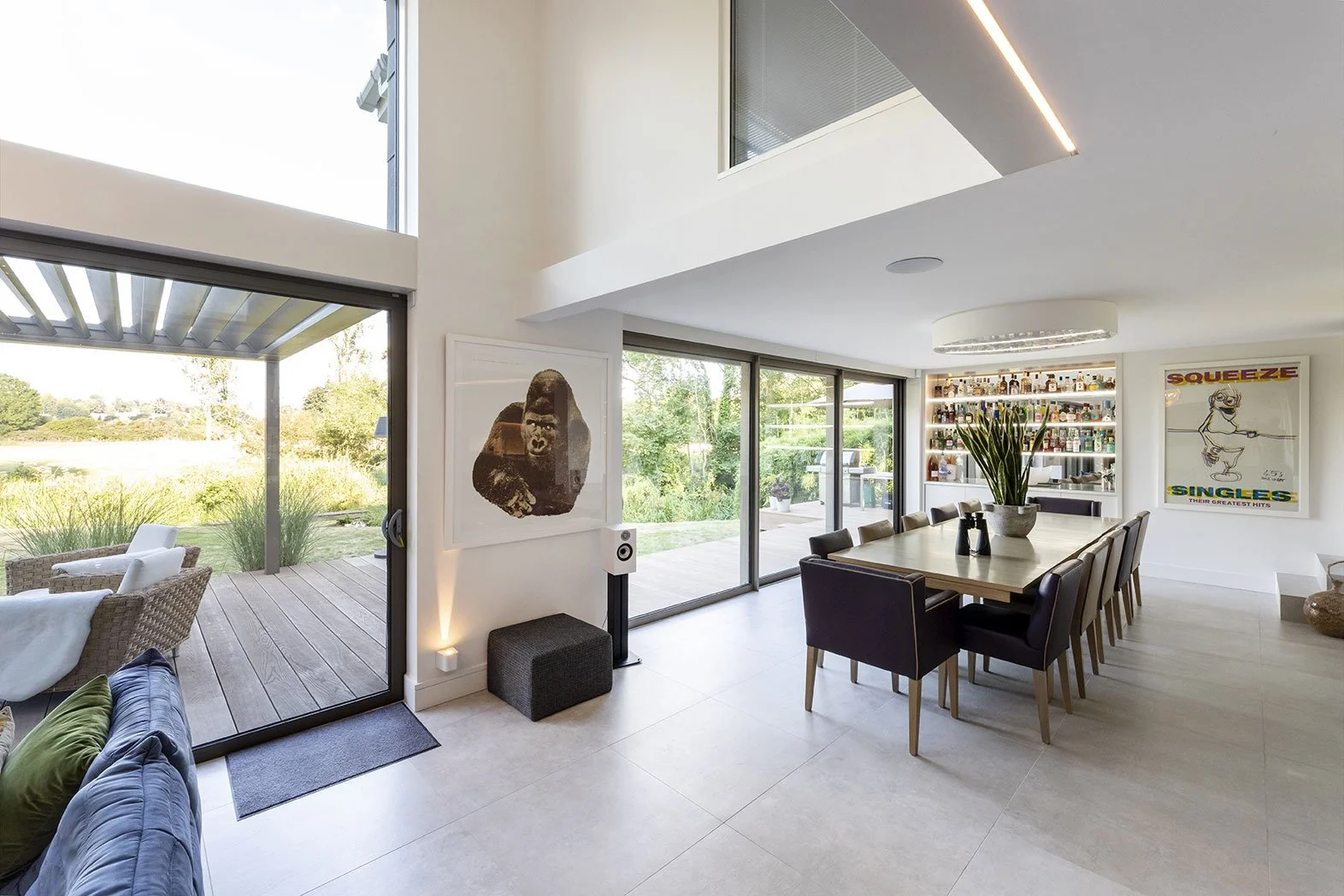 Modern dining room with a large wooden dining table, black chairs, and a bar area in the background. The room has large sliding glass doors that lead to a deck and outside greenery. There are contemporary artworks on the walls, a potted plant on the 