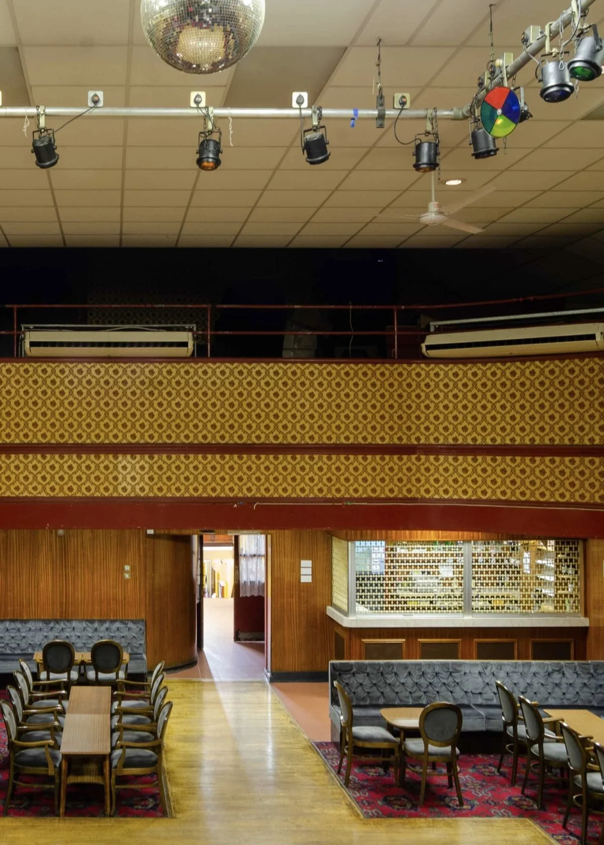 Interior of a vintage restaurant or cafe with wooden walls, patterned golden walls, and vintage carpeted seating areas. There is a stage or performance area with stage lights and equipment hanging from the ceiling.