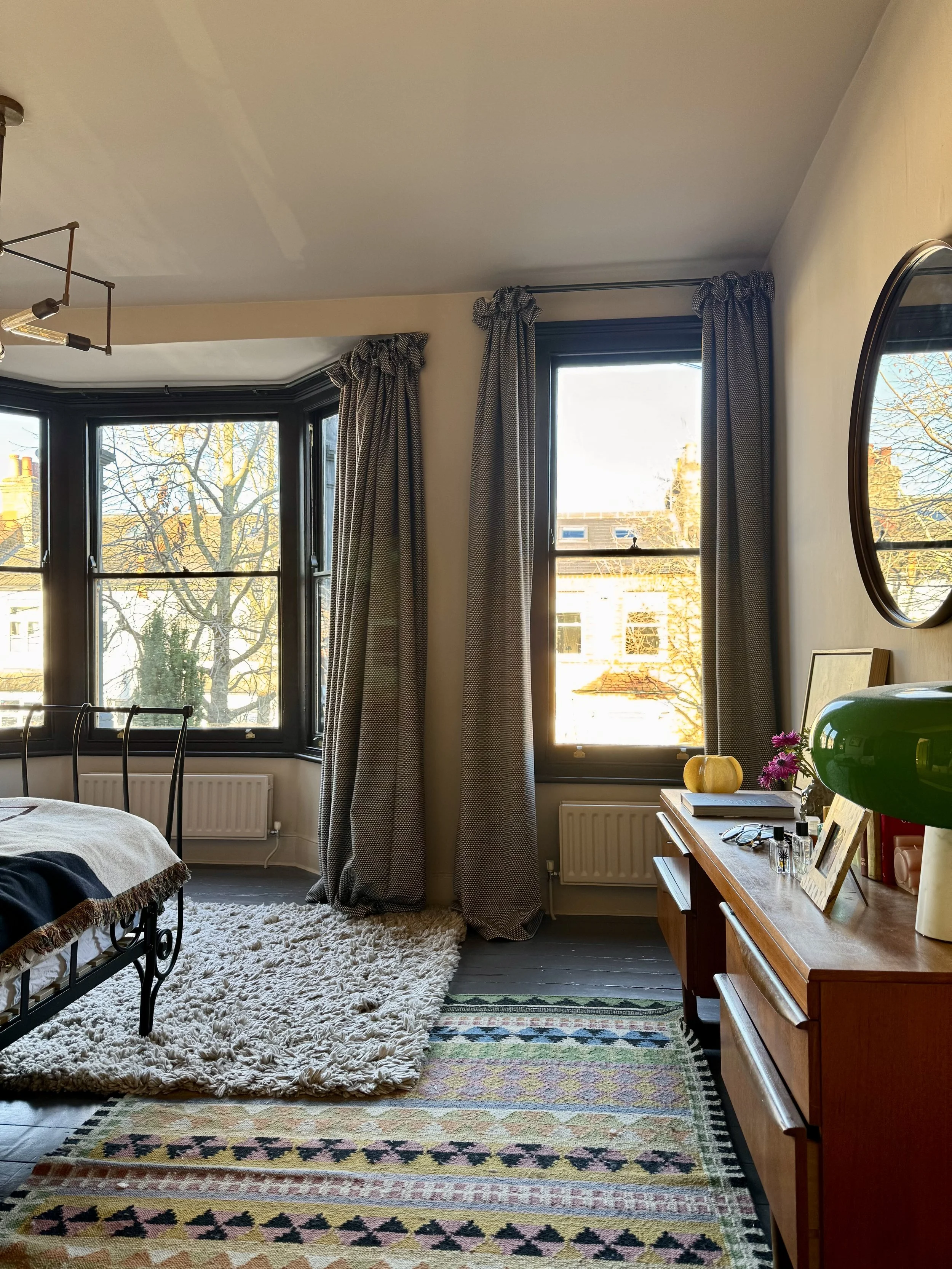 Sunlit bedroom with two large windows, patterned curtains, a wooden dresser with decorative items, colorful rug, and a bed with a blanket.