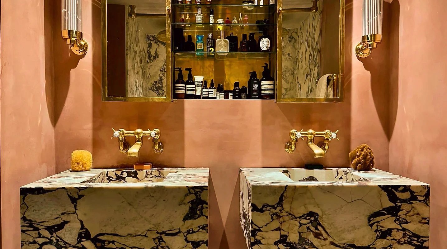 Dual marble sinks with gold faucets in a luxurious bathroom, with pink walls, gold mirrors, and a mirrored cabinet storing toiletries and beauty products.