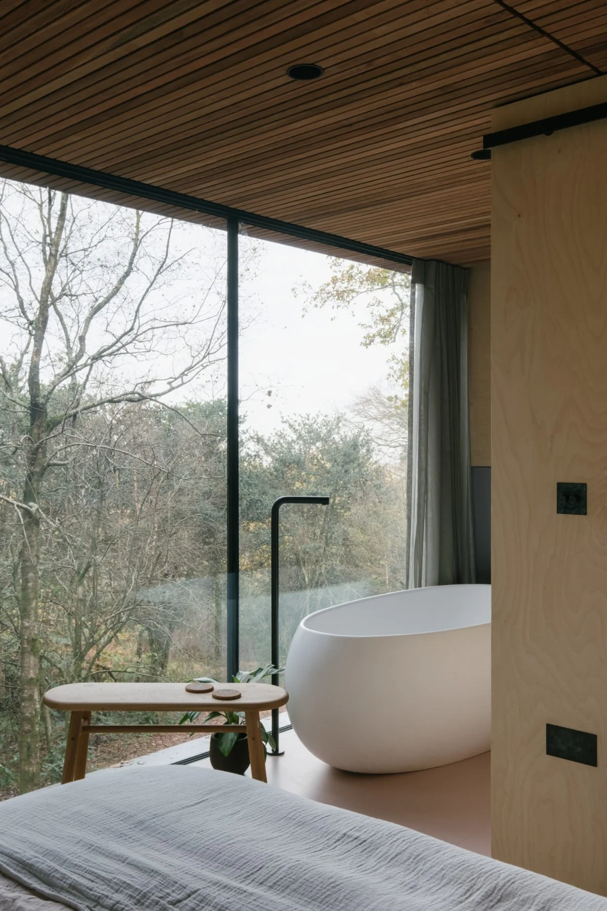 Modern bathroom with a freestanding bathtub near a large glass window overlooking trees, small wooden side table, and a bed in the foreground.