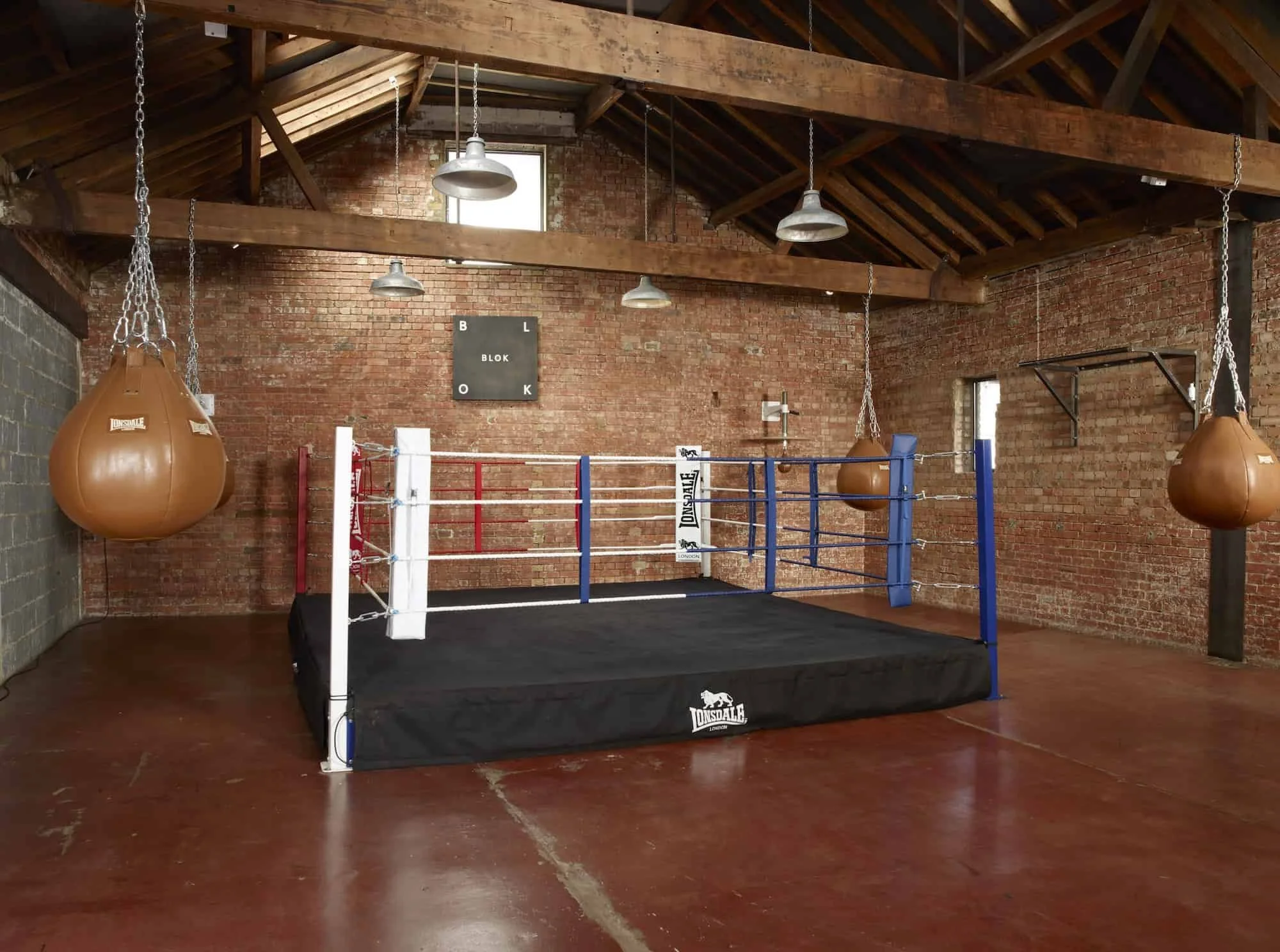 Boxing gym with brick walls and wooden ceiling, featuring a boxing ring with red, white, and blue ropes, two brown punching bags hanging from the ceiling, and industrial-style lighting fixtures.
