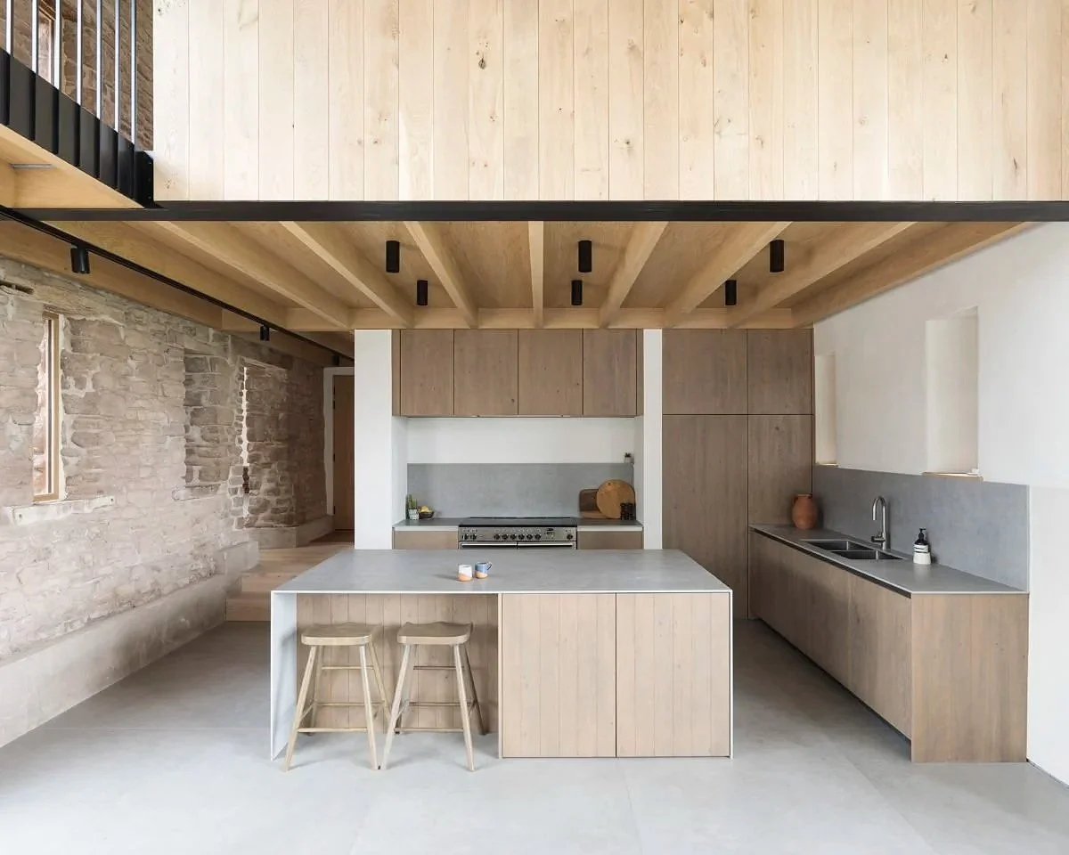Modern kitchen with wooden cabinets, a stone wall, and a large island with a gray countertop, two wooden stools, and minimal decorations.