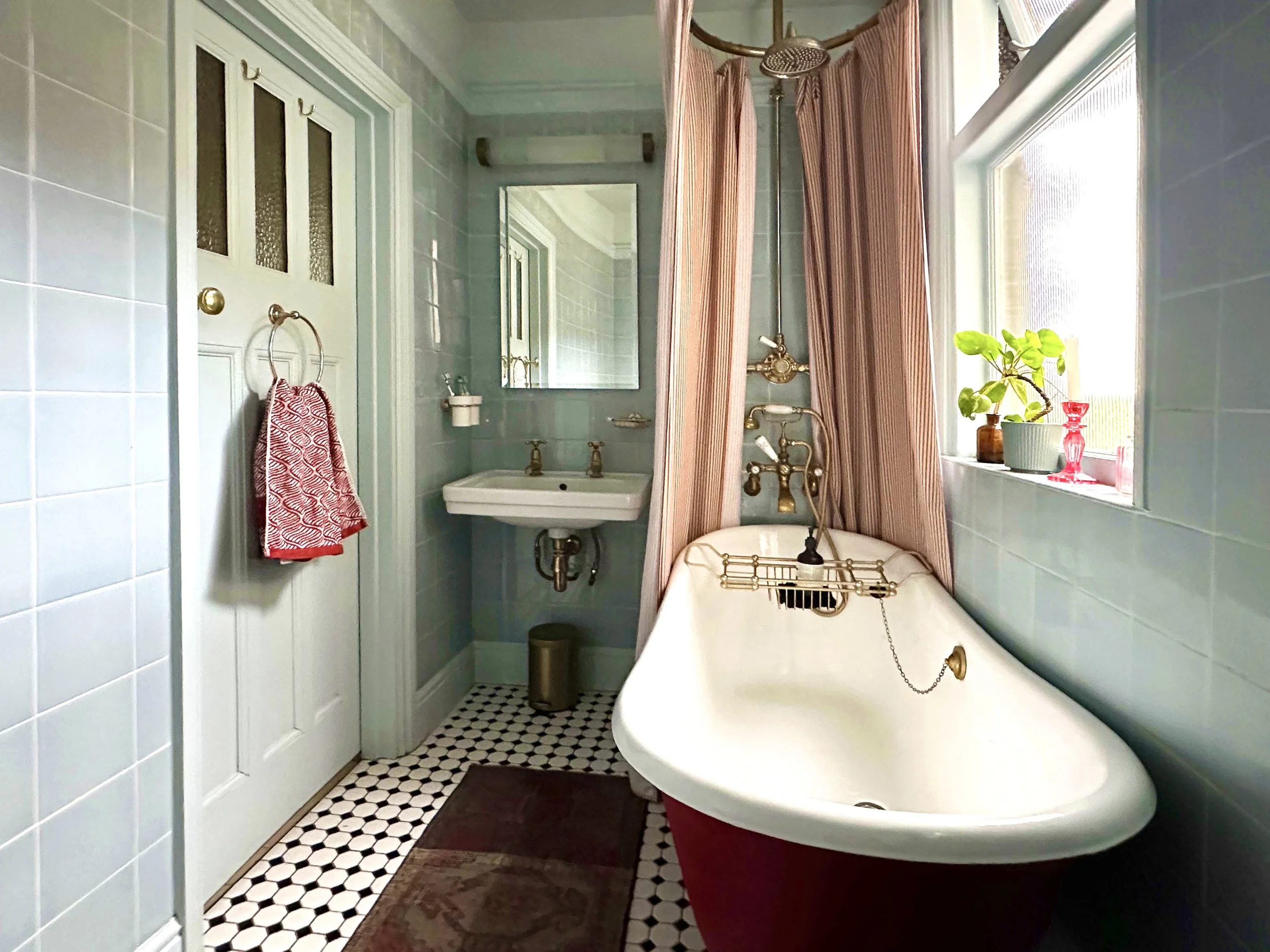 Vintage bathroom with a clawfoot bathtub, pink and white striped shower curtain, small sink with brass fixtures, mirror, potted plants on the windowsill, and patterned tile floor.